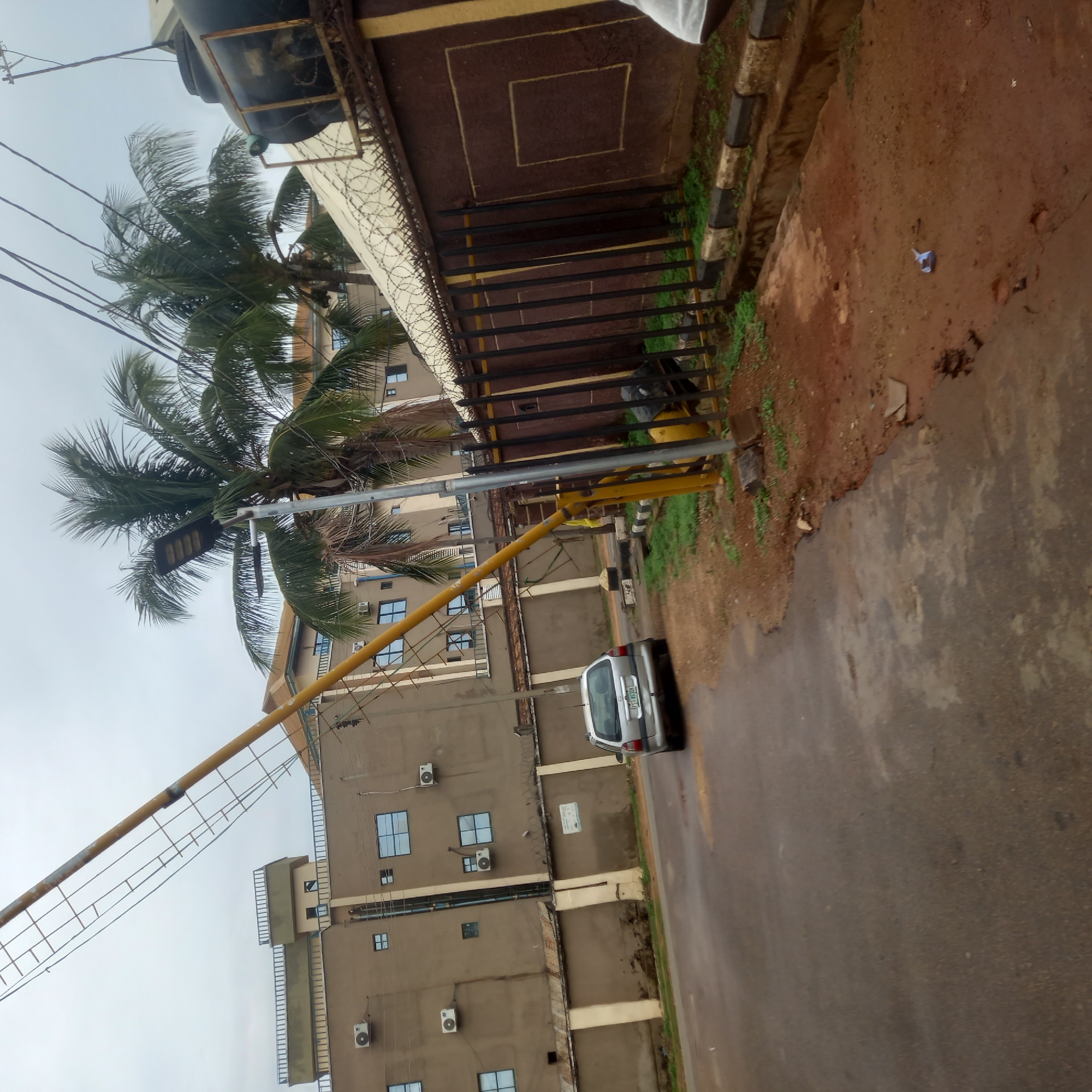 The image shows a street scene in Barden Yana, Nigeria. A silver car is parked on the right side of the street, next to a yellow security gate. Behind the gate is a tall, beige building with multiple windows and air conditioning units. A palm tree is partially visible in the foreground. Above the building is a brown wall topped with barbed wire. The sky is overcast. The ground beside the road is a mix of brown soil and some sparse green vegetation.