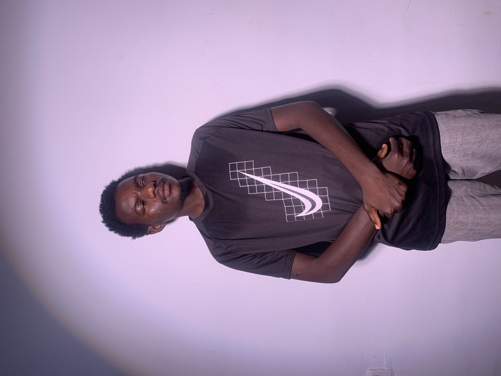A young Black man with short, dark, curly hair and a goatee is lying on his side, facing the camera. He is wearing a black t-shirt with a white Nike logo and a geometric design on the chest, and light gray sweatpants. His left arm is bent and resting on his chest, with his hand clasped over his right arm. The man's eyes are closed, and his mouth is slightly open, giving him a relaxed or pensive expression. He is positioned against a plain, light-colored wall, with a distinct shadow cast to his right, indicating a light source from the left and slightly above. The lighting creates a soft gradient across the background, transitioning from a bright, illuminated area on the left to a darker, shadowed area on the right. The image has a slightly tilted perspective. No text is visible other than the logo on the shirt.