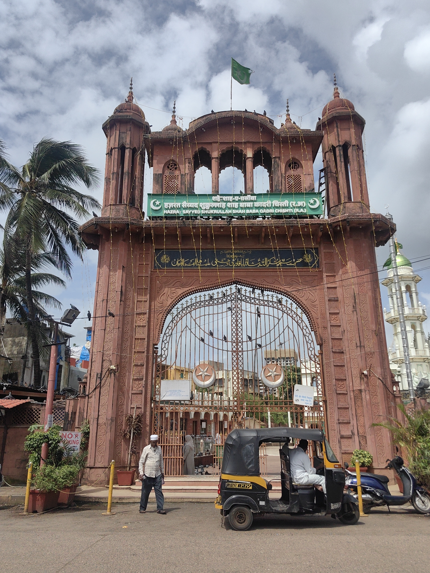 The image depicts the entrance to a large, ornate building with red sandstone architecture. The building features prominent minarets and decorative arches, suggesting it is a religious or historical landmark. A green flag with a white crescent moon and star is visible flying from the top of the structure. Below the flag, a green banner displays text in both Hindi and English, identifying the location as "Hazrat Sayyed Shukrullah Shah Baba Qadri Chishti (R.A.)". This indicates the building is likely a shrine or mosque dedicated to this individual, located in Mumbai, India.
In the foreground, an auto-rickshaw is parked on the right side of the road. A man wearing a white shirt and dark pants walks towards the entrance on the left side of the image. Another auto-rickshaw, predominantly black and yellow, is partially visible on the right, with a person seated inside. A blue scooter is parked behind the auto-rickshaw.
The environment suggests a street scene in Mumbai, with visible palm trees on the left, and other buildings and a white minaret of a mosque in the background. The sky is partly cloudy. Decorative string lights are hung across the entrance archway, indicating a festive occasion or ongoing decoration. A "NO PARKING" sign is visible near the plants on the left.