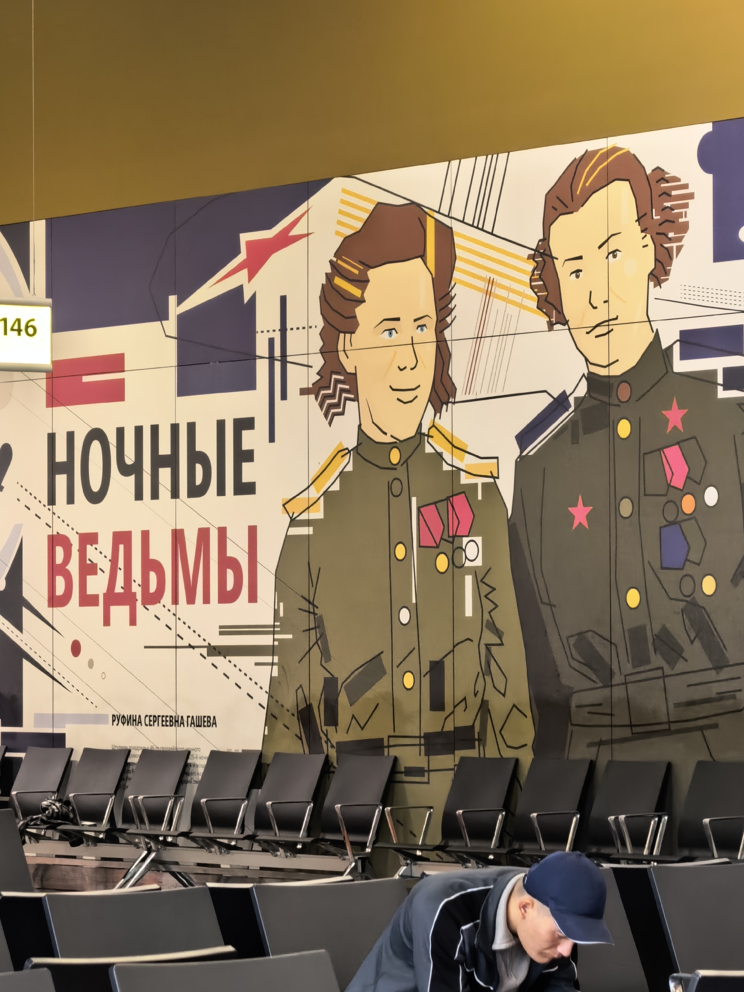 The image shows a mural on a wall featuring two stylized figures in military uniforms. The mural is vibrant with bold lines and colors. Below the mural, there is a row of empty black seats arranged in a waiting area, possibly an airport or train station. A person wearing a dark jacket and a blue cap is bent over in the foreground, out of focus. Text in Cyrillic script is visible on the wall, with large letters spelling "НОЧНЫЕ ВЕДЬМЫ" (Night Witches). Another line of text identifies "РУФИНА СЕРГЕЕВНА ГАШЕВА". A sign with the number "146" is also visible on the left side of the image.