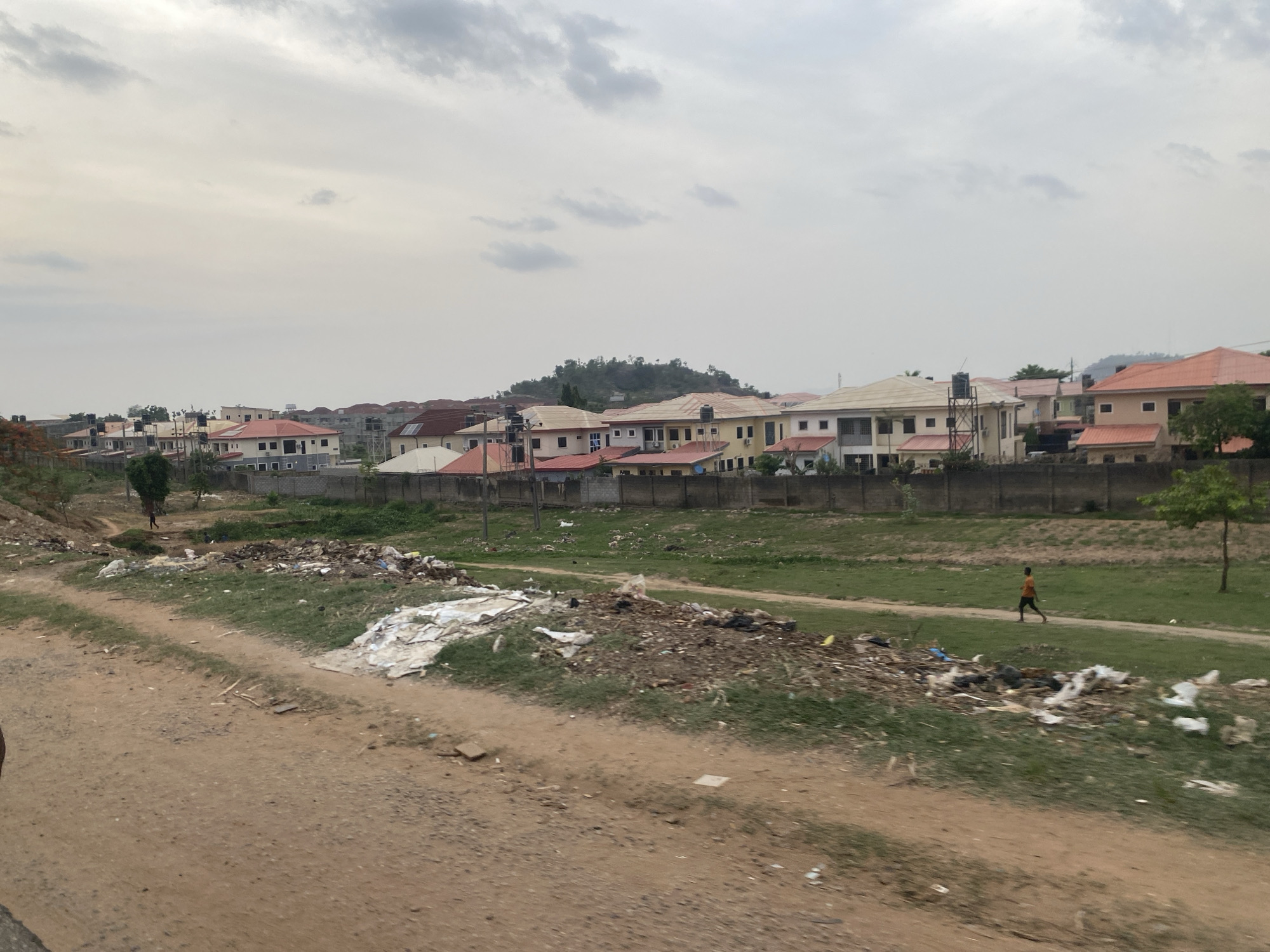 The scene is an impoverished area on the outskirts of Abuja, Nigeria. A dirt road lines the foreground, with large piles of trash scattered along its edge. Beyond the trash, a narrow strip of green grass separates the road from a gray concrete wall, behind which stand several single-story residential buildings with red and cream-colored roofs. A small, tree-covered hill rises in the background. A young boy in an orange shirt is walking along the grassy strip. In the distance, another person is visible near a tree. The sky is overcast.