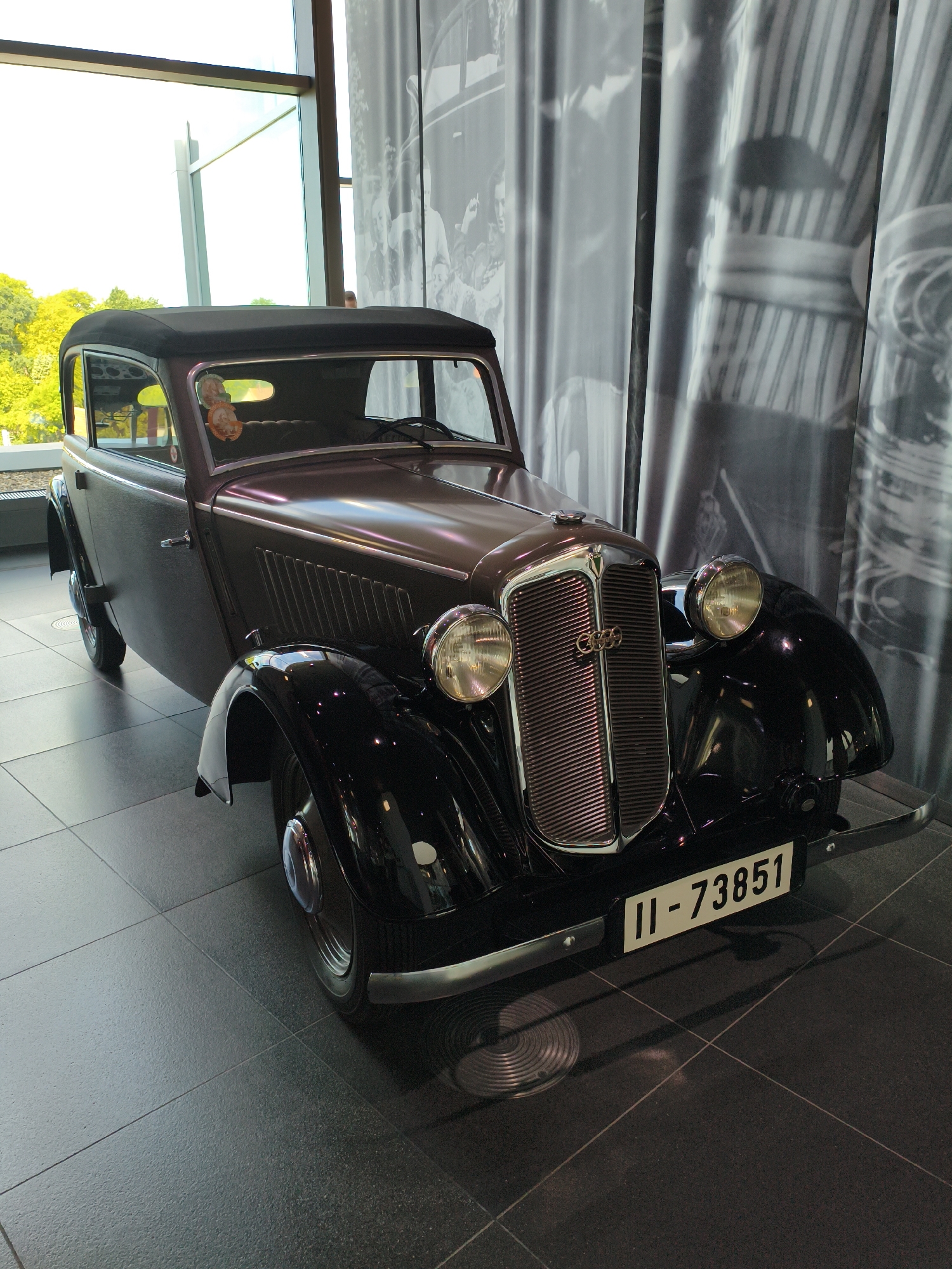 A black vintage Audi sedan is parked on a tiled floor inside a modern building. The car has a convertible top and chrome accents on the grille, headlights, and bumper. A license plate with the number "II-73851" is affixed to the front bumper. The interior of the car is visible through the windows, and a pair of decorative ornaments hang from the rearview mirror. Behind the car, a large black and white mural depicts people and architectural elements, with vertical fabric panels obscuring parts of the mural. To the left of the car, a large window reveals an outdoor scene with green trees and a bright sky. This scene is likely located at the Audi Forum in Ingolstadt, Germany, a museum dedicated to the history of Audi automobiles. There are no people visible in the immediate vicinity of the car.