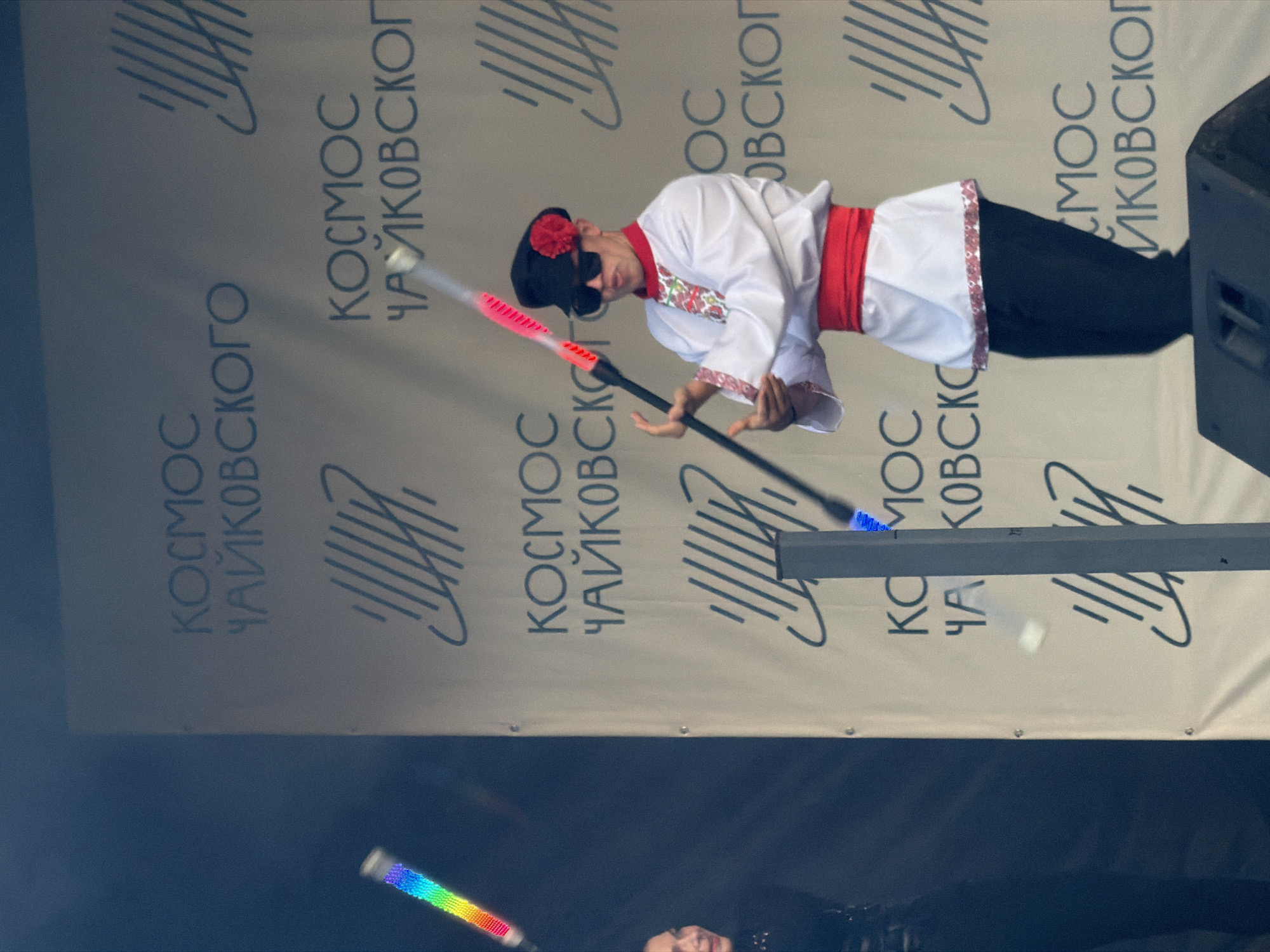 An outdoor daytime performance is taking place in front of a large banner that reads "КОСМОС ЧАЙКОВСКОГО" in Russian, indicating the event is related to the city of Chaikovsky, Russia. A male performer, dressed in a traditional white shirt with red embroidery and a red sash, black pants, a black cap adorned with a red flower, and sunglasses, is juggling two illuminated props. One prop, with a rainbow-colored light, is held to his left, while the other, glowing with red light, is in the process of being juggled. To the left of the performer, a portion of another person's head and shoulder is visible, also illuminated by a prop. The stage appears to be a large speaker or sound system.