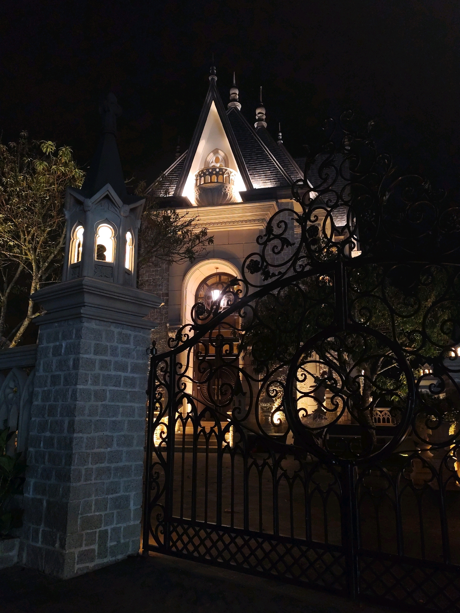 The image shows a nighttime view of the entrance to Dinh 1, also known as the Palace of the Governor-General of Indochina, in Da Lat, Vietnam. A large, ornate black iron gate, featuring intricate scrollwork and Gothic-inspired arched elements, stands in the foreground. Beyond the gate, a stone pillar with a small, illuminated Gothic-style structure is visible to the left. The main building, Dinh 1, is illuminated from within and by external lights, highlighting its colonial French architecture with a pitched roof, pointed spires, and arched windows. Trees with bare branches are present on the grounds. The overall scene conveys a sense of historical grandeur and nighttime ambiance.