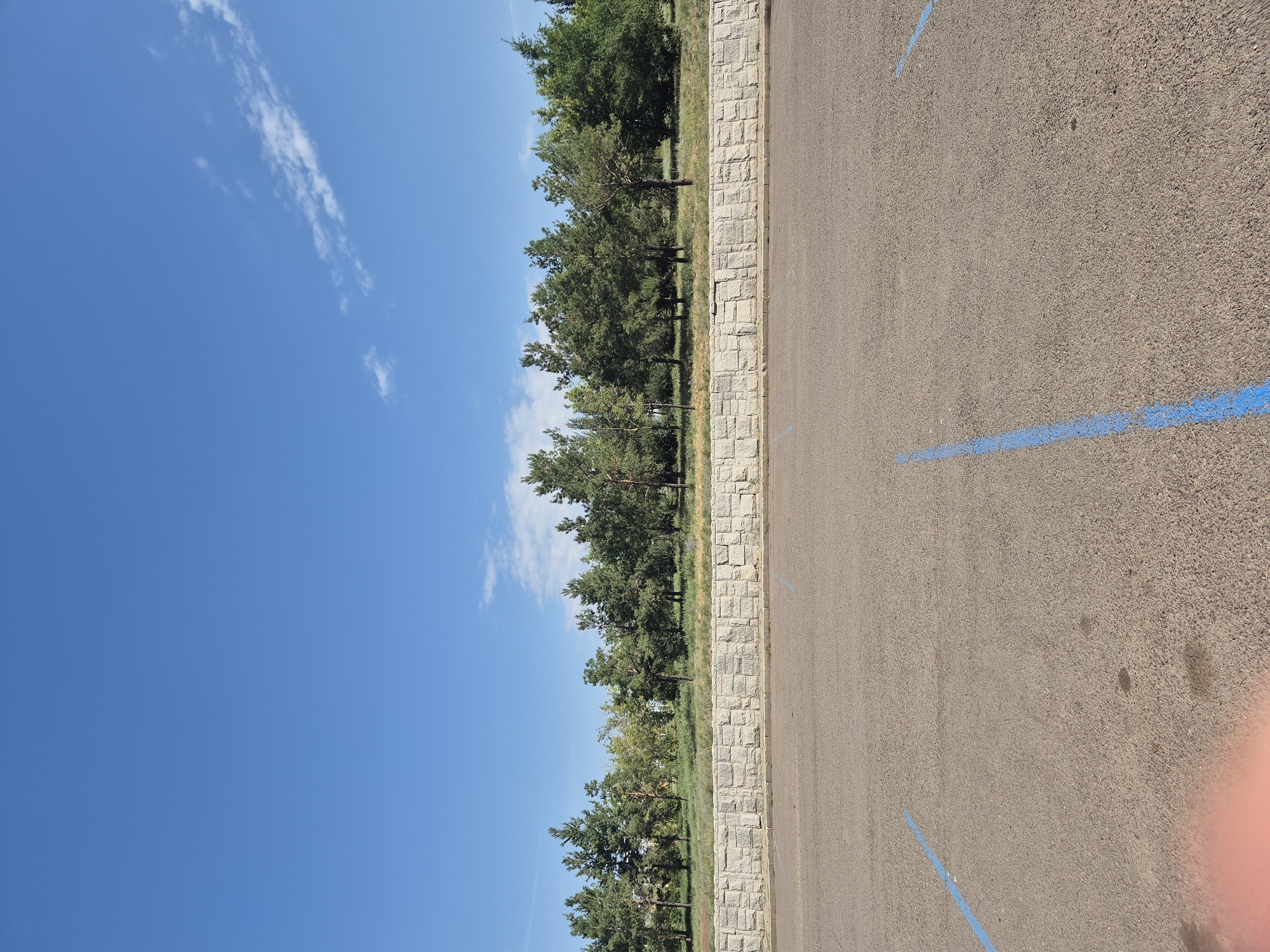 A full shot of an empty parking lot in Astana, Kazakhstan, under a bright blue sky. The parking lot is asphalt, with faint blue painted lines marking parking spots. In the background, a stone retaining wall separates the parking lot from a grassy area dotted with various trees. The trees are a mix of evergreen and deciduous species, with lush green foliage. The sky is clear with a few wispy white clouds. The image appears to be taken during the daytime.