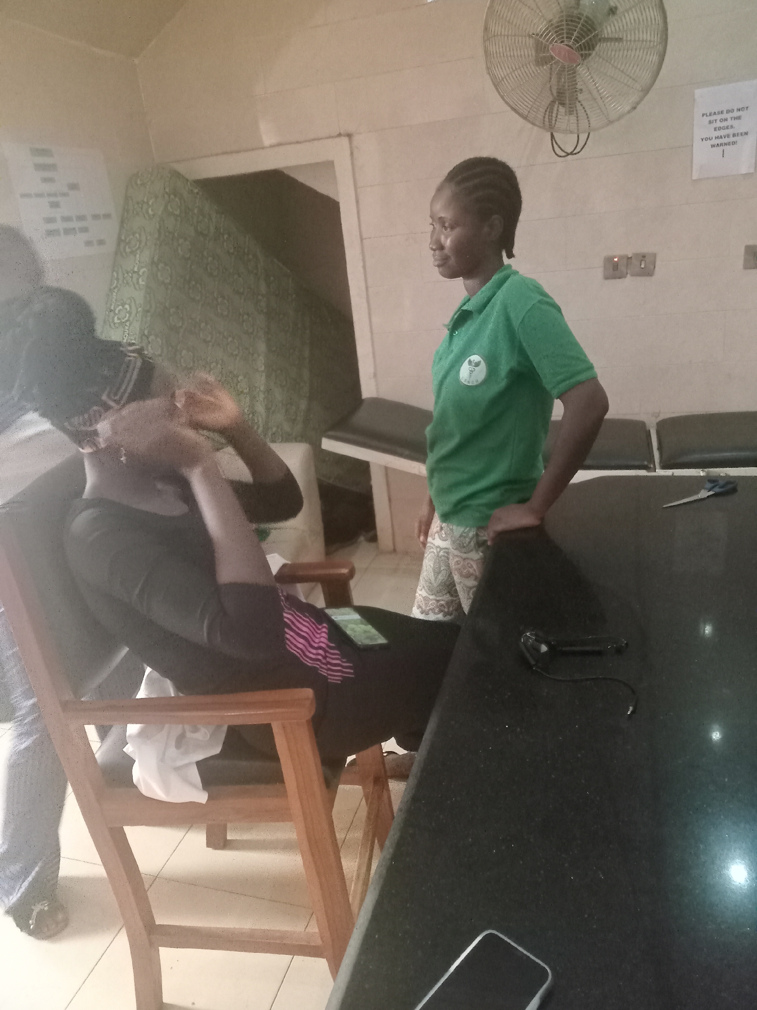 The image depicts two individuals in what appears to be a medical or clinic setting, possibly in Gwarinpa, Nigeria, given the instruction. One person is seated in a wooden chair, wearing dark clothing with a pink stripe, and their hands are near their face. The second person is standing, wearing a green shirt and patterned shorts, with their hand resting on a dark, possibly granite countertop. On the countertop are a pair of scissors, a small electronic device, and a smartphone. A wall fan is visible in the background along with a sign that reads, "PLEASE DO NOT SIT ON THE EDGES. YOU HAVE BEEN WARNED!" There's also a padded bench/examination table near the back. Another individual is seen to the left, wearing jeans, partially out of view.