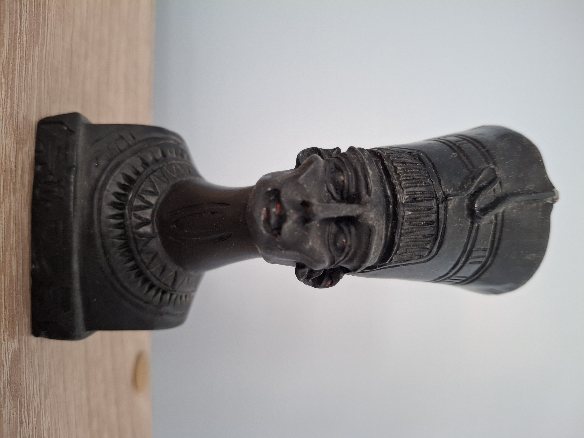 A dark-colored statue of an Egyptian pharaoh head and neck rests on a wooden surface. The background is a gradient of white to light blue. The statue's facial features are visible, including the nose, lips, and headdress. The statue's base has a circular, textured design. The statue is located in Pleven, Bulgaria.