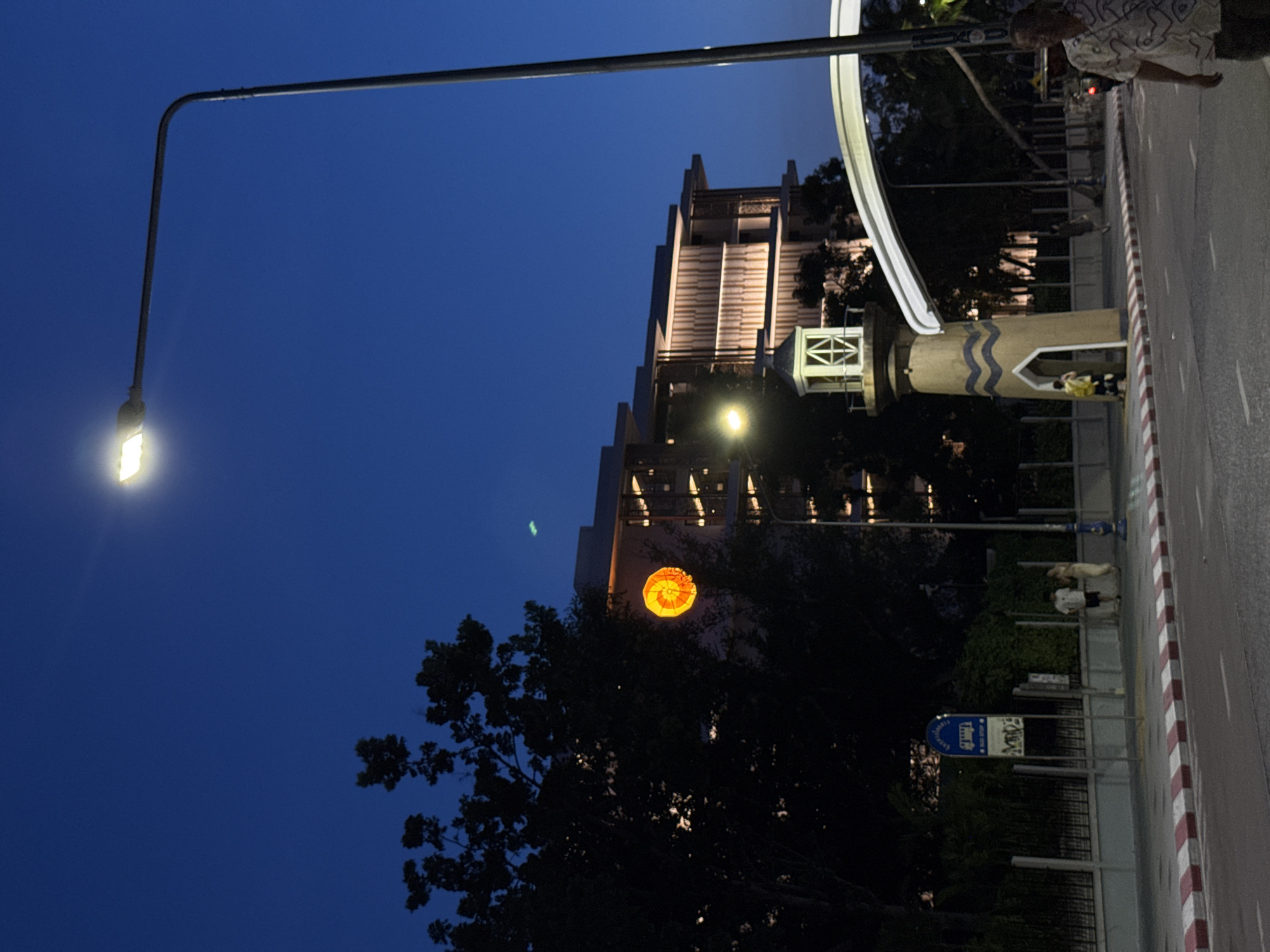 At dusk, a street scene unfolds in Pattaya City, Thailand. A tall, modern streetlamp with a bright LED bulb illuminates the left side of the frame. To the right, a multi-story building with contemporary architecture is visible, its facade lit with horizontal stripes of warm light. In the foreground, a stylized lighthouse-like structure stands near the road, featuring a beige base with blue wave patterns and a white, lantern-like top. A white archway curves over the road behind the lighthouse. The sidewalk is bordered by a curb with alternating red and white stripes. Two people are walking on the sidewalk near a bus stop sign. On the road, a person is walking towards the camera, wearing a patterned shirt. Trees frame the background, silhouetted against the deep blue twilight sky.