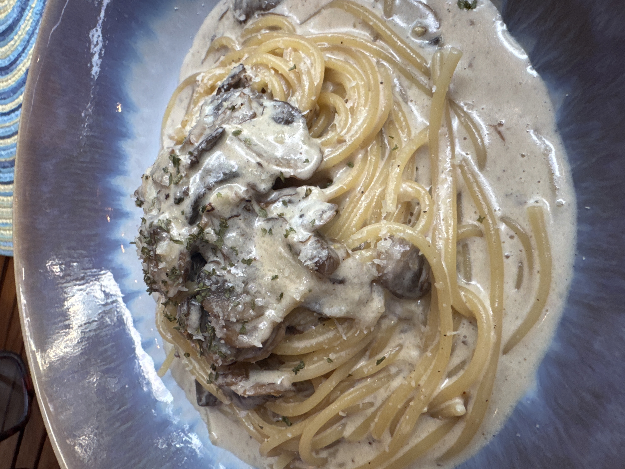 A close-up, high-angle shot shows a plate of creamy mushroom pasta. The pasta is light yellow and coated in a white, creamy sauce. A generous portion of mushrooms, also covered in the sauce, is piled on top of the pasta. Small green herbs are sprinkled over the mushrooms. The plate is blue with white marbling effects. A sliver of a striped blue and white textile is visible in the upper left corner. The plate appears to be sitting on a dark wooden surface. This scene is believed to be located in Baan Mae Nam, Thailand.