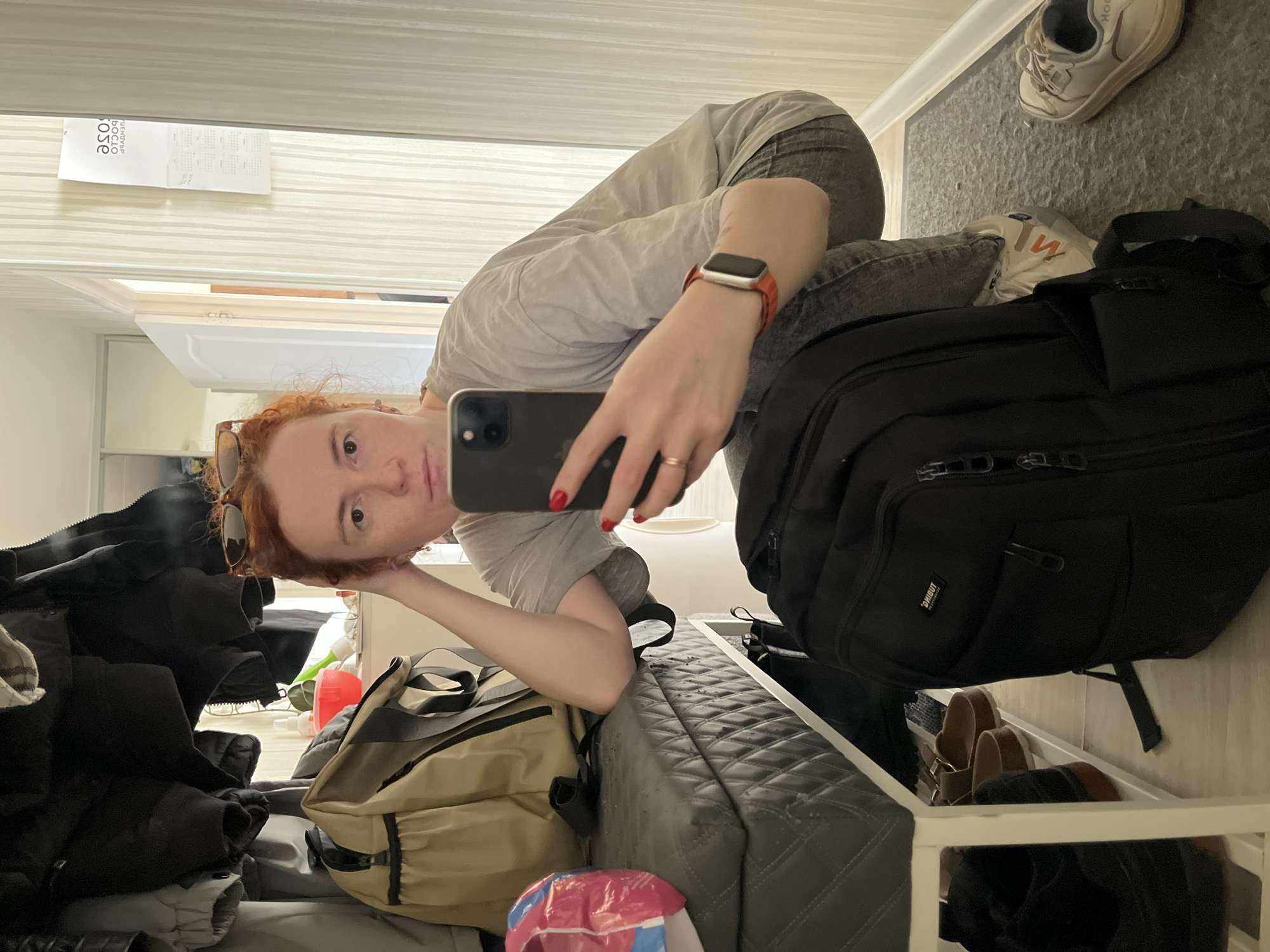 A selfie taken in an interior space, possibly a hotel room in Chaikovsky, Russia based on the calendar hanging on the wall that says "Чайковский" in Russian. A person with red hair is holding a smartphone and taking the picture, wearing a grey t-shirt and grey jeans. They are also wearing an Apple Watch with an orange band. The room appears cluttered. To the person's right is a large black backpack and a pair of white sneakers on the floor. Behind the person, there are clothes piled up. The room has a door visible in the background. Also visible is a tan backpack. Underneath what appears to be a suitcase are three pairs of shoes.