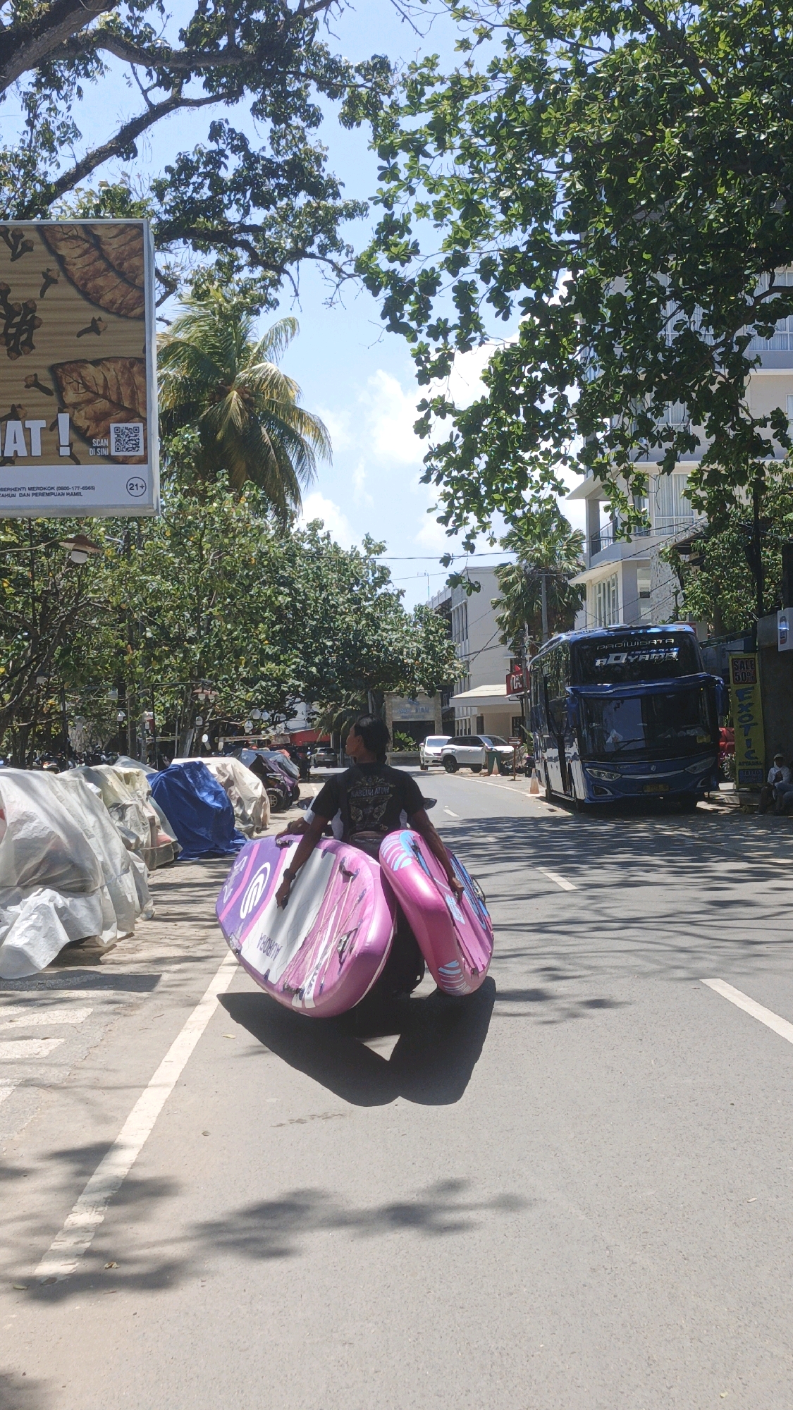 In Desa Pagandaran, Indonesia, on a sunny day, a person walks down a street carrying two inflatable paddleboards, one pink and one purple with pink accents. The person, seen from the back, wears a dark t-shirt with lettering on it and dark pants. They are walking on the right side of the road, facing away from the camera. The paddleboards are stacked and held by the person.
The street is paved with asphalt and has a white line marking the lane. To the left of the person, there are several objects covered with tarps, possibly parked vehicles or stored items. Beyond these, trees and foliage line the sidewalk. In the background, a large blue tour bus with "PADI WISATA" visible on its side is parked on the right side of the road. Further back, there are multi-story buildings, some with balconies. Green trees with lush foliage are abundant throughout the scene, casting shadows on the street. The sky is bright blue with scattered white clouds. The overall environment appears to be a commercial or tourist area.