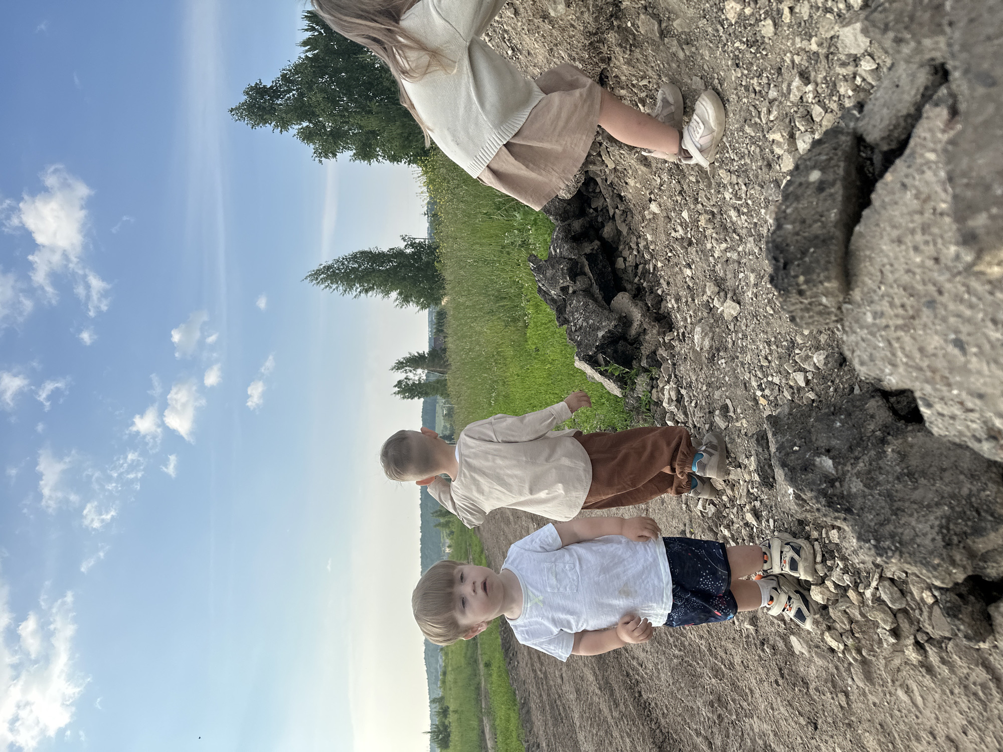 Three children are on a dirt path near a grassy field under a partly cloudy blue sky. In the foreground to the left, a young boy with light brown hair is looking slightly to his left. He is wearing a white t-shirt, dark blue shorts with a white pattern, and light-colored sneakers. Behind him and to the right, another young boy with light brown hair is facing away from the camera, looking towards the right. He is wearing a light beige long-sleeved shirt and brown pants, with light-colored sneakers. To the far right, a child with long blonde hair is leaning forward and looking down, partially obscured by a pile of rocks. This child is wearing a light beige sweater and a light brown skirt, with light-colored sneakers. The environment appears rural, with tall grass and trees visible in the background. The path is composed of dirt and gravel, with scattered rocks and some broken asphalt pieces in the foreground. There is no indication of the city of Estachi, Russia in the image.