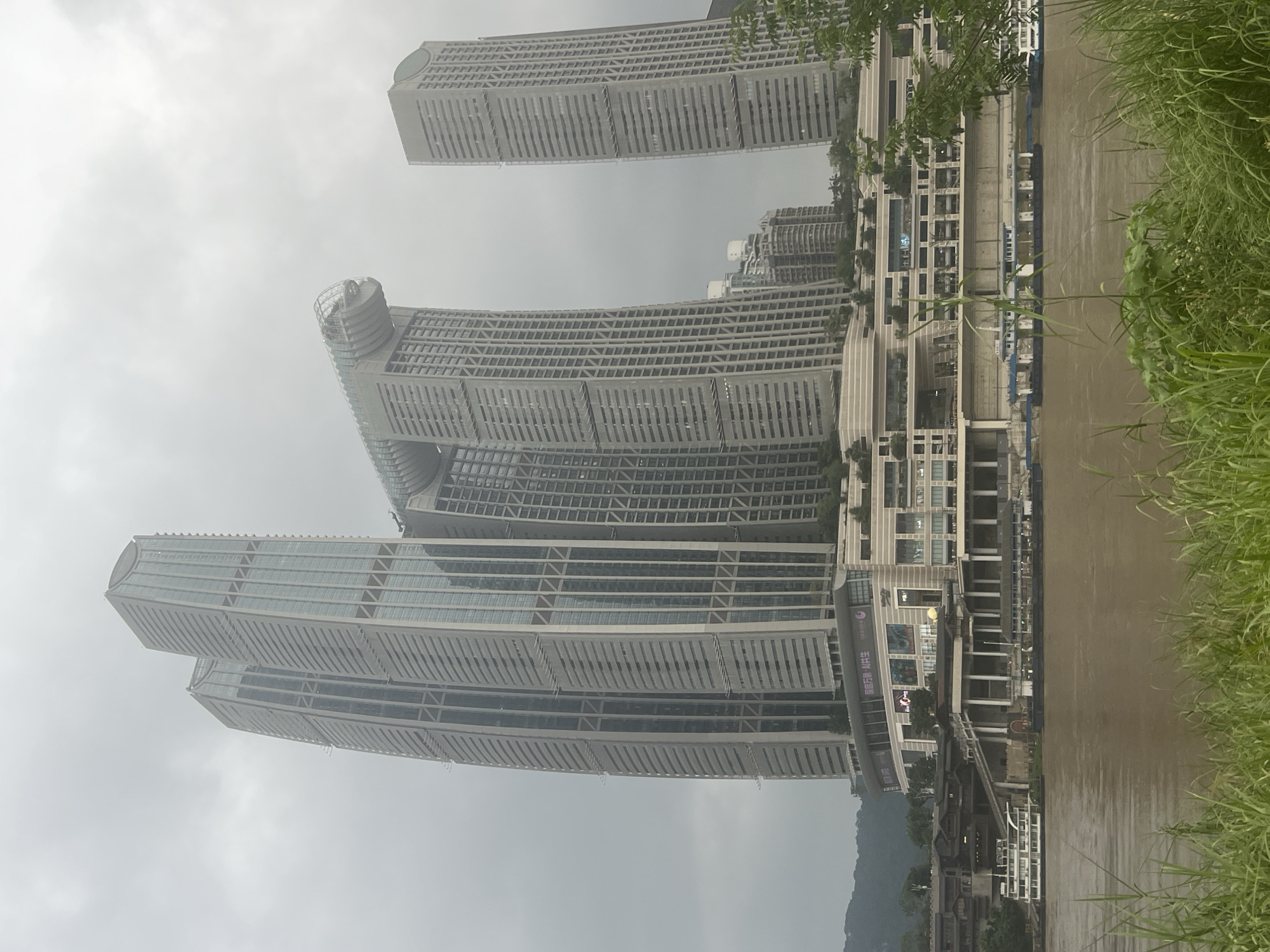 The image depicts the Raffles City Chongqing complex, a mixed-use development located in the Yuzhong District of Chongqing, China. The complex is situated on the confluence of the Yangtze and Jialing Rivers, with the muddy brown water of the Yangtze visible in the foreground. Several boats, including a white ferry and a blue and white passenger boat, are docked or navigating the river.
The architectural centerpiece of the complex is a striking cluster of skyscrapers, dominated by two unusually shaped towers that are connected by a horizontal structure resembling a ship. The towers have a modern, geometric design, with a facade of glass and grey panels. To the right of these main towers stands another tall, rectangular skyscraper. Below the skyscrapers is a multi-tiered commercial and residential area, featuring shops, restaurants, and open spaces with greenery. The sky is overcast and grey, contributing to a muted and somewhat somber atmosphere. Lush green grass and foliage are visible along the riverbank in the foreground, contrasting with the man-made structures. No people are discernible in the image.