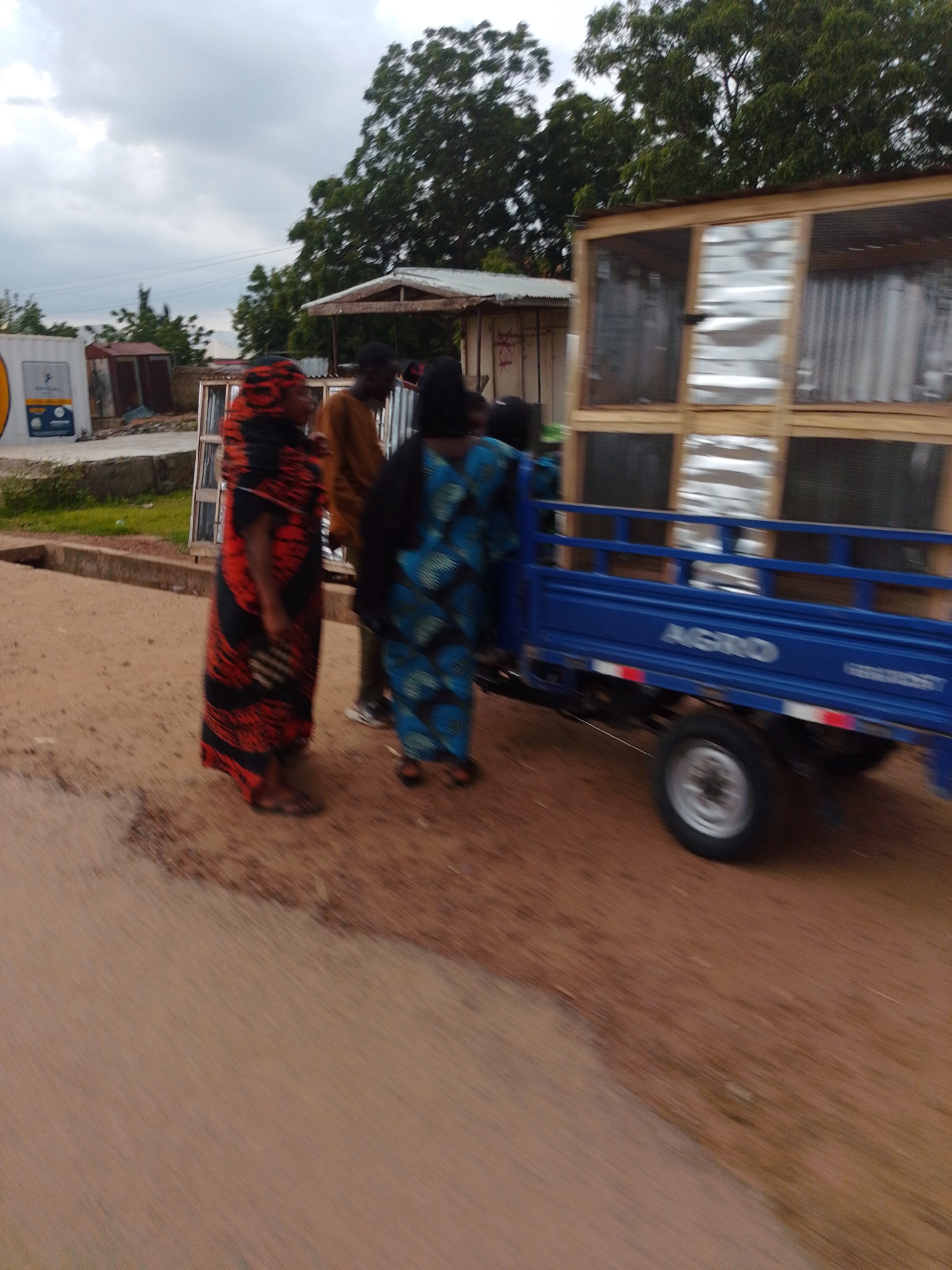 In the city of Gombe, Nigeria, an outdoor scene unfolds on a dusty road. In the foreground, a woman dressed in a vibrant red and black patterned garment stands to the left, her form partially obscured by motion blur. Beside her, a man in a brown top faces away from the camera, his features indistinct due to the blur. Another woman, wearing a blue and black patterned dress, is positioned to the right of the man.
Behind the individuals is a small blue truck, its "AGRO" branding visible on the side, also affected by motion blur. The truck's bed contains a wooden structure with metal sheeting, resembling a cage or enclosure. The environment is characterized by dry, reddish-brown earth and sparse vegetation, with trees visible in the background under a cloudy sky. In the far left of the image, a white structure with a yellow circular emblem and some text is partially visible, suggesting a commercial or administrative building. The overall impression is one of a busy street scene, possibly a market or a transport hub, captured during a moment of movement.