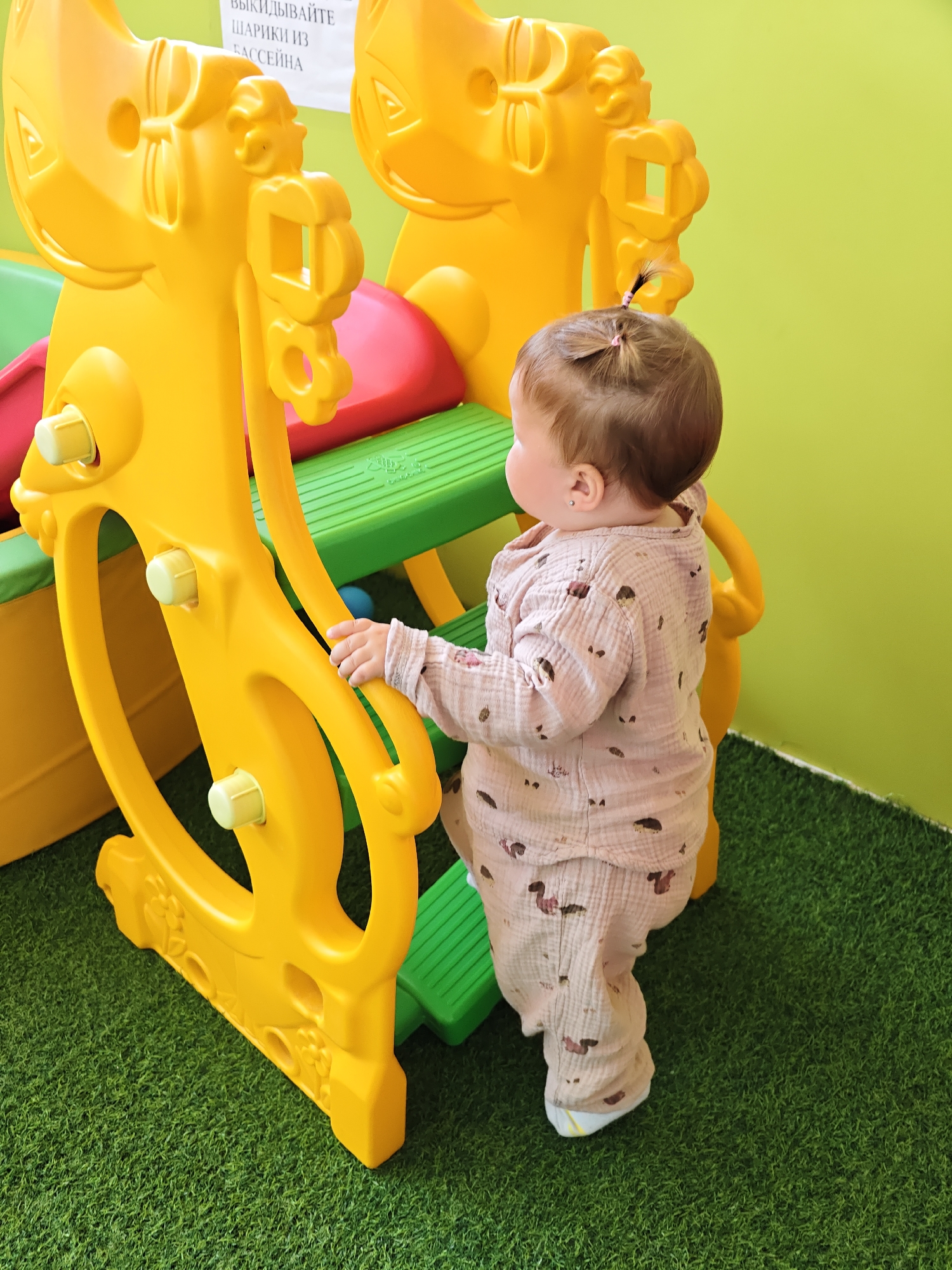 A toddler with light brown hair tied in two small pigtails, wearing a light pink gauze pajama set with a subtle pattern, stands on green artificial turf in front of a bright yellow plastic playground structure. The toddler is facing away from the camera, looking towards the playground, and has their right hand on a yellow support beam of the structure. The playground has red and green plastic components. A sign with Russian text is visible on the wall in the background, which is a light green color. The scene appears to be indoors, possibly in a play area within the city of Chaikovsky, Russia, given the provided context.