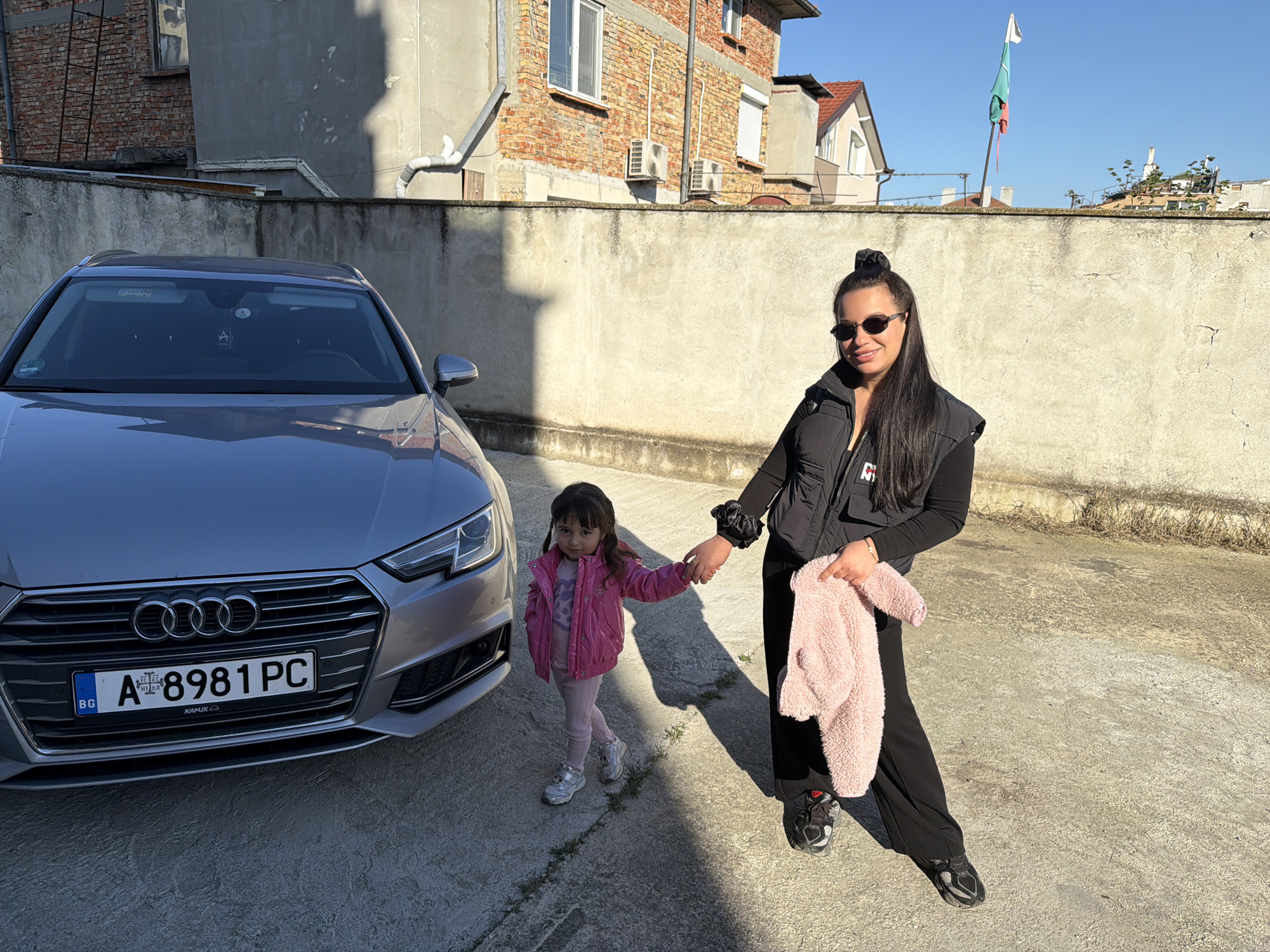 The photograph depicts a woman and a young girl standing on a concrete surface next to a gray Audi A4 car. The woman, wearing sunglasses, a black vest, and black pants, holds the girl's hand and a fluffy pink garment. The girl is dressed in a pink jacket and leggings. The car is positioned near a concrete wall, with buildings and a Bulgarian flag visible in the background. The scene is bathed in sunlight. The license plate of the car reads "A 8981 PC". The location is Unknown, Unknown.