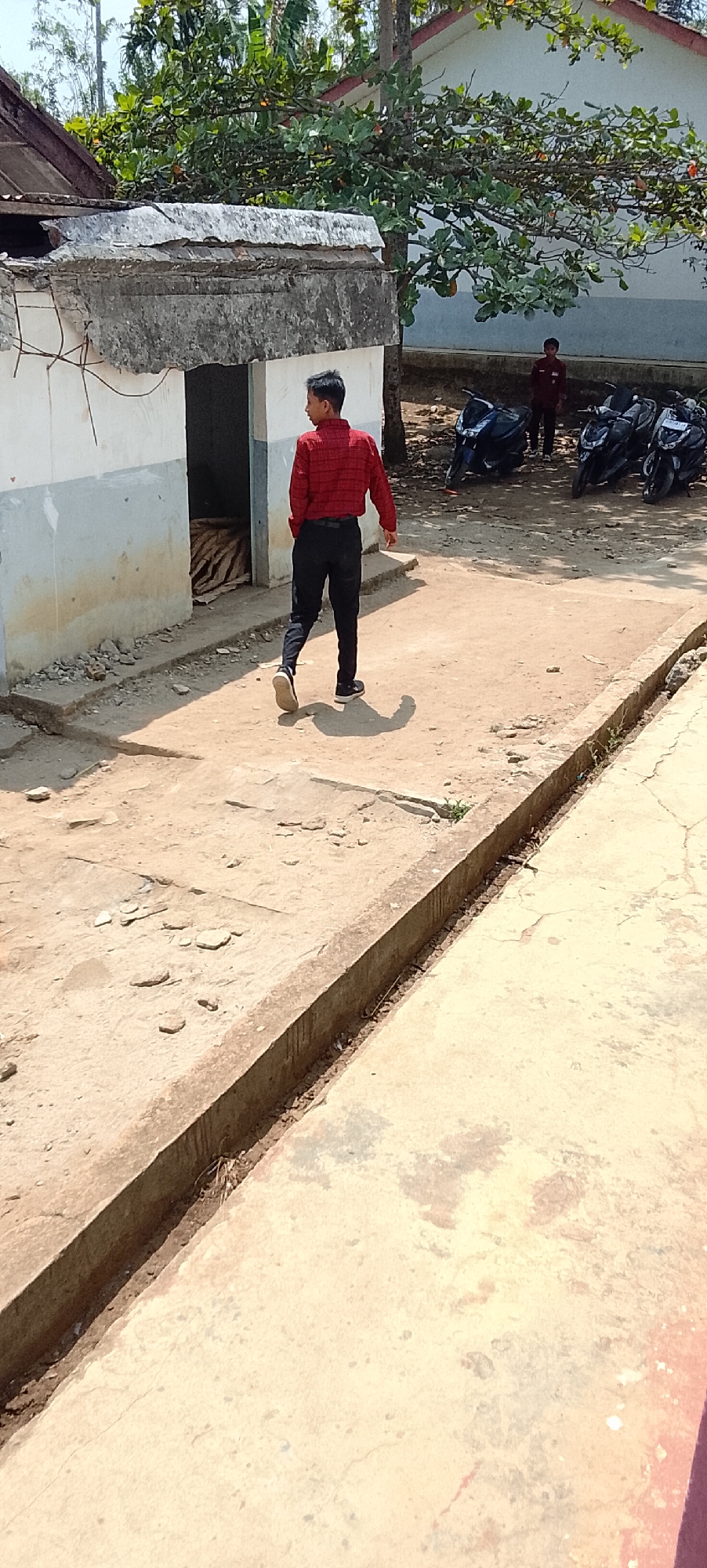 A young man in a red checkered shirt and black pants walks away from the camera on a dirt and concrete path outside a dilapidated building. The building has a broken concrete overhang. To the right of the building are several parked motorcycles. In the background, another young man stands near the motorcycles. The environment is outdoors with trees and vegetation visible. The scene is lit by bright sunlight. The location is identified as Tanjung Setia, Indonesia.
