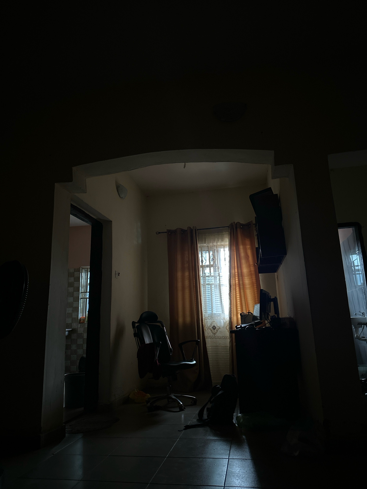 The image depicts an indoor scene, likely a room in a residential or office setting, in Umuahia, Nigeria. The lighting is dim, with light filtering in from a window covered by orange curtains. The window itself has a white patterned sheer curtain behind the orange one. In the foreground, a dark office chair with a headrest is positioned. To the right of the chair, a dark-colored cabinet or desk is visible, upon which a dark object, possibly a laptop or monitor, is placed. A black backpack is on the floor in front of the cabinet. To the left, an open doorway reveals a glimpse of another room, where a window with tiled walls and a sink can be seen. The flooring appears to be tiled. The overall impression is of a dimly lit interior space with furniture and an entrance to another area.