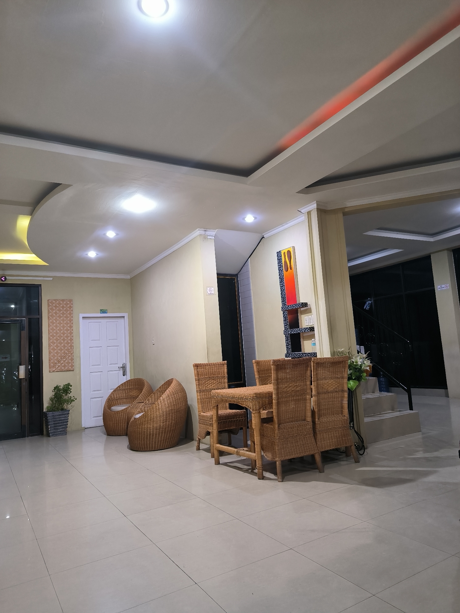 The image captures an unoccupied indoor common area, likely a hotel lobby or guesthouse reception, illuminated by artificial lighting, indicating evening or night. The floor consists of large, light-colored square tiles.
In the foreground and midground, a collection of light brown wicker furniture includes a dining table with four chairs and two curved lounge chairs. A potted plant in a grey planter sits near a glass door, which reveals a dark exterior. A white paneled door is marked with the number "105" above it, and a woven decorative panel hangs on the adjacent wall.
Further into the room, an abstract artwork featuring reds, oranges, and blues is mounted on the wall above the wicker table. Steps lead upwards on the right side of the frame, next to which a small white floral arrangement is placed. The walls are painted a light cream color.
The ceiling features multiple recessed circular lights, some intensely bright, and a distinct red strip light embedded within a drop ceiling structure. A "NO SMOKING" sign is visible on a pillar to the right. The setting appears calm and ready for guests, consistent with a facility in Banda Aceh, Indonesia.