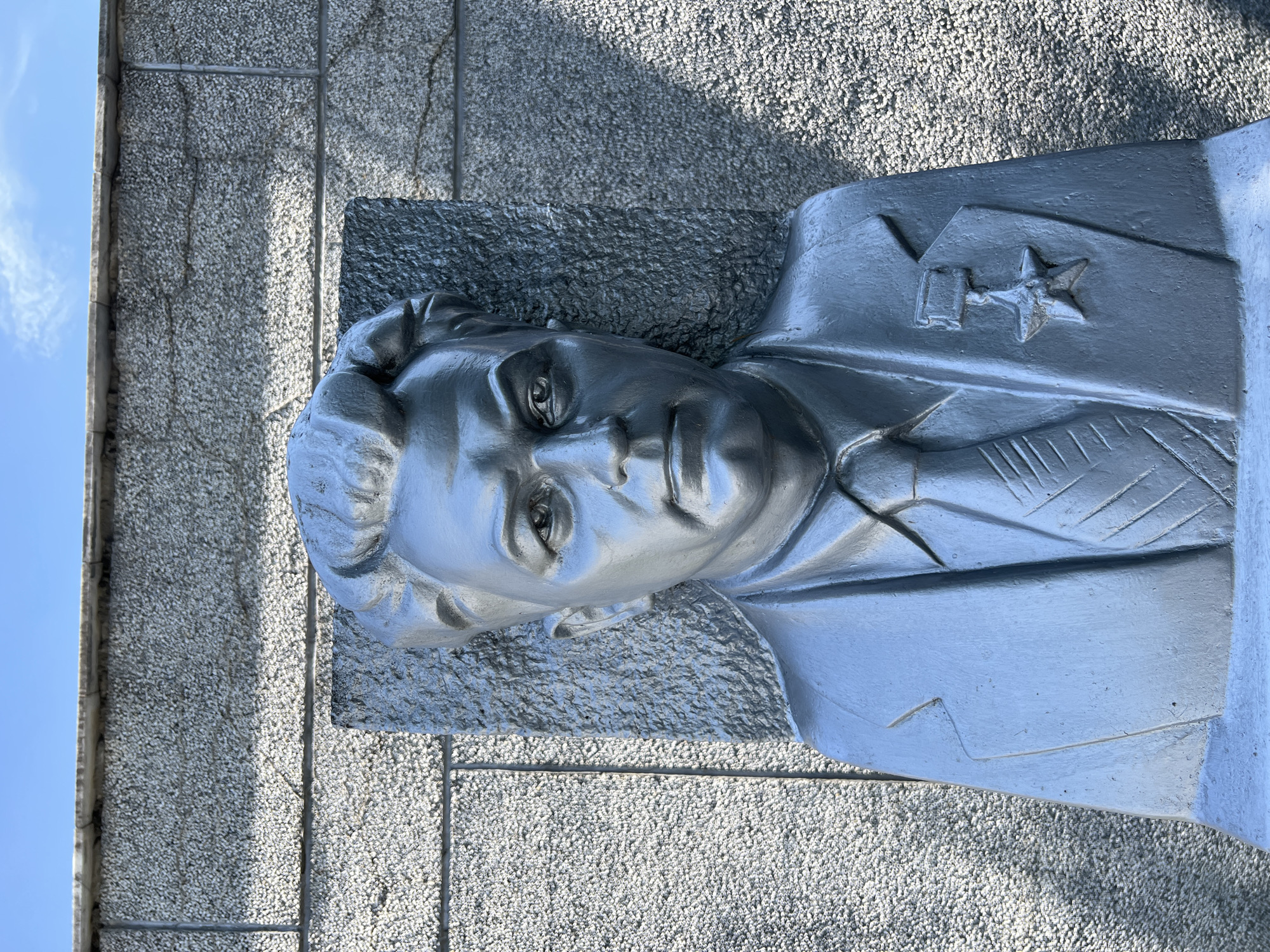 The image shows a close-up, rotated view of a silver-colored bas-relief sculpture of a man's head and torso. The sculpture is set against a textured, grey stone wall, with a bright blue sky visible in the upper left corner. Sunlight casts shadows on the wall, suggesting it is daytime. The man in the sculpture has a stern expression, with detailed features such as the eyes, nose, and mouth. He is depicted wearing a uniform with what appears to be a medal or badge on his chest, including a star and a rectangular emblem. The texture of the stone wall is coarse and speckled, with visible grout lines separating the blocks. The overall impression is one of a memorial or monument. Given the location context of Takhtamukai, Russia, this sculpture likely depicts a historical figure or a military hero significant to the region. The lighting and crisp details suggest a clear, possibly sunny day.
