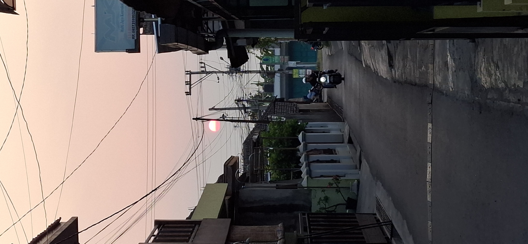 The image depicts a narrow street in Wonorejo, Indonesia, at sunset. The sky is a soft pinkish-orange, and the sun is a visible red orb near the horizon, partially obscured by power lines. The street is lined with houses and shops, many with visible electrical wiring and utility poles. A person on a motorcycle, wearing a helmet, is riding away from the camera down the street. Another person is visible on a motorcycle further down the road. There are speed bumps on the asphalt road. The overall atmosphere is that of a residential or commercial street in a densely populated area during the transition from day to night.