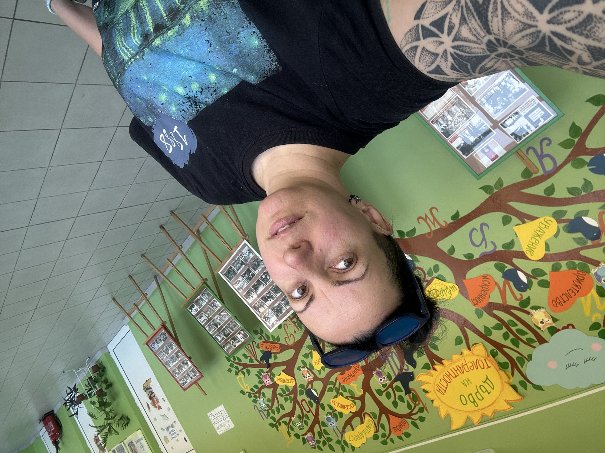 The image shows a woman in an indoor space, likely a school or community center, in Radomir, Bulgaria, with a wall decorated with a large tree mural. The mural features a tree with owls, leaves, and labels in Bulgarian, promoting values like "friendship," "respect," "sincerity," "gratitude," and "tolerance." She is wearing a black shirt with an inscription that reads "1883." She is also wearing sunglasses and has tattoos on her right arm. The walls are green, and framed photos are displayed. The floor is tiled in neutral tones.