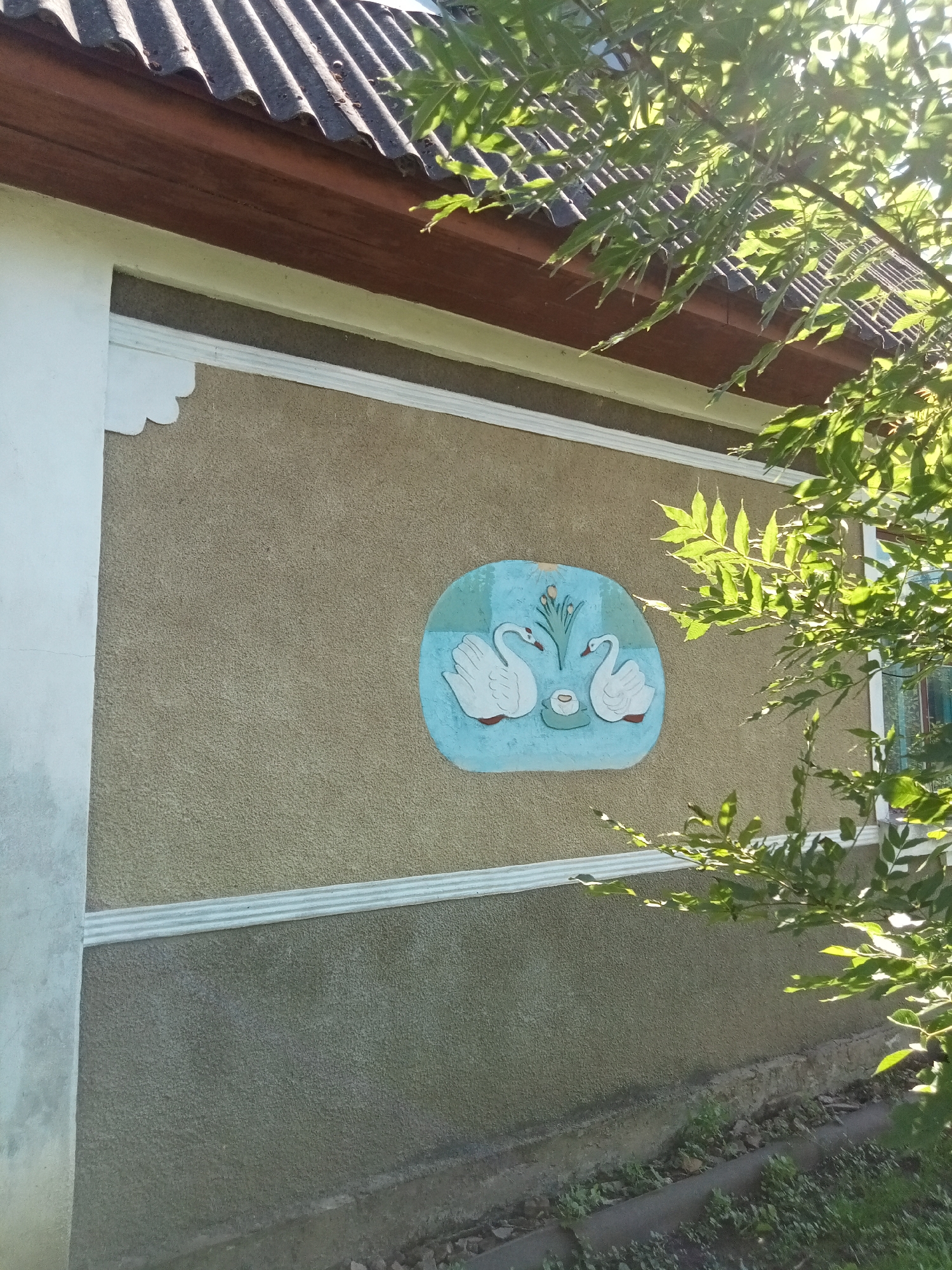The image shows a close-up view of the textured wall of a building, possibly a house. The wall is painted a beige or tan color and is adorned with a decorative mural. The mural is set within an oval frame and depicts two white swans facing each other, with a small dish and some yellow flowers between them. The swans appear to be on a blue body of water, suggested by the background of the mural.
Above the wall, the corrugated roof of the building is visible, supported by a dark brown wooden fascia. A white decorative trim runs horizontally along the wall, dividing the upper and lower sections. On the left side of the wall, a section of white painted trim is visible, possibly part of an architectural detail.
The scene is bathed in sunlight, indicated by the bright illumination and the dappled shadows cast by the leaves of a tree in the foreground. The leaves are lush and green, suggesting it is daytime and likely spring or summer. The sunlight filtering through the leaves creates a natural, somewhat diffused lighting effect.
The overall impression is one of a rustic or traditional dwelling, with the painted mural adding a touch of artistic flair. The setting appears to be outdoors, given the presence of the tree and the natural light. There is no visible text in the image.