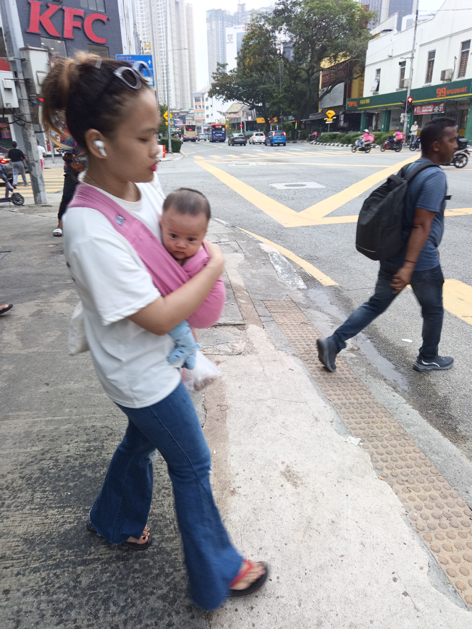 A woman is walking on a sidewalk next to a street in Kuala Lumpur, Malaysia, with a baby strapped to her chest. The woman is wearing a white t-shirt, blue bell-bottom jeans, and brown sandals. She has sunglasses perched on her head and is wearing white earbuds. The baby is swaddled in a pink sling and is wearing blue socks. The background shows a busy city street with buildings, vehicles, and pedestrians. A KFC restaurant is visible on the left side of the image. A man with a backpack is walking on the street on the right side of the image. The sky is overcast.