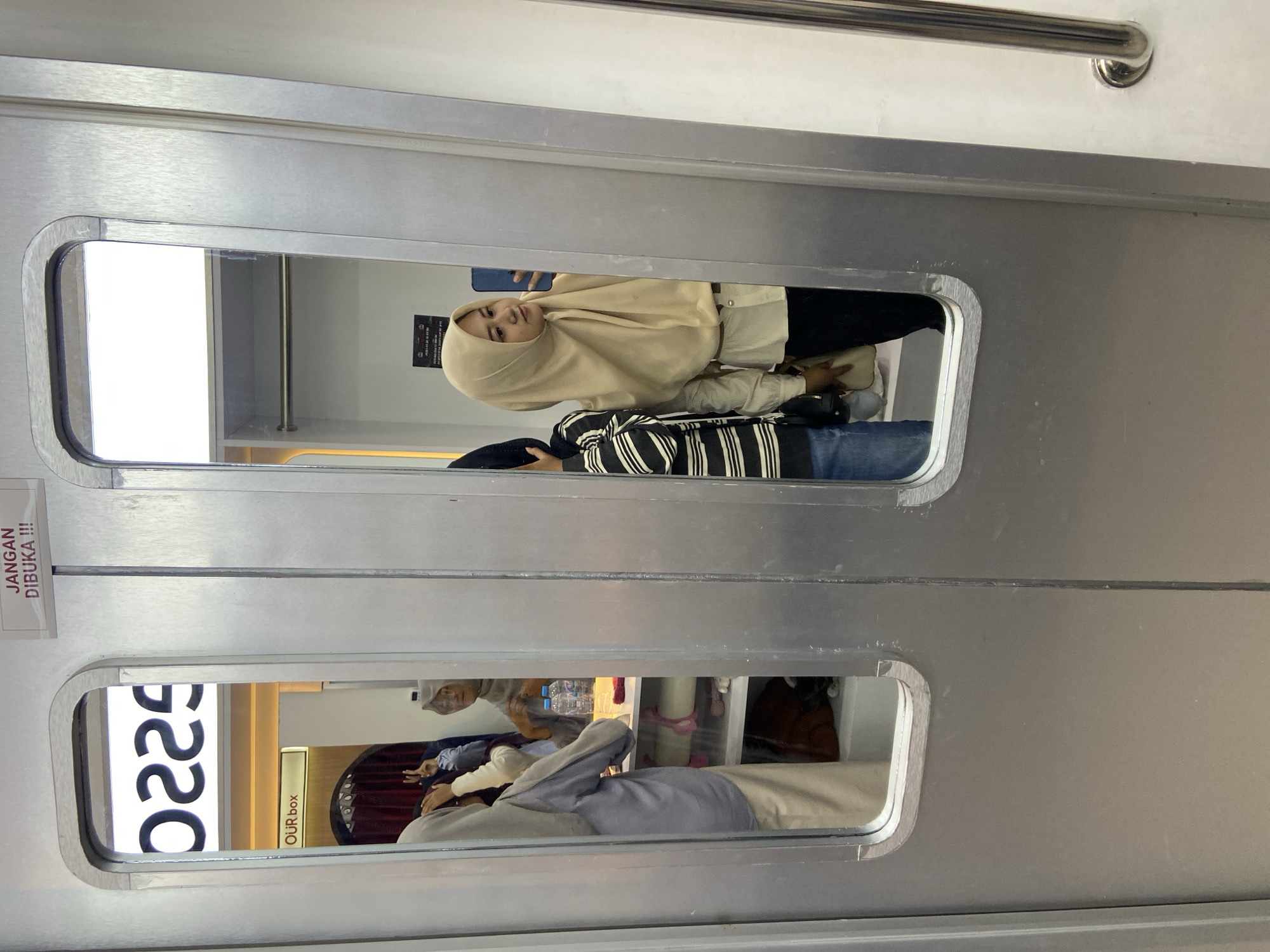 The image shows a view through two rectangular windows on a train car. The windows look into separate compartments. In the top window, a woman wearing a hijab is taking a selfie. She is standing with another person, who is wearing a striped shirt and jeans. The lower window shows a group of people sitting in a compartment that appears to be a small dining area or lounge, as indicated by the text "OUR BOX". A large white sign with black numbers, "0225," is visible. The scene is likely on a train in Sidoarjo, Indonesia, based on the location tag provided.