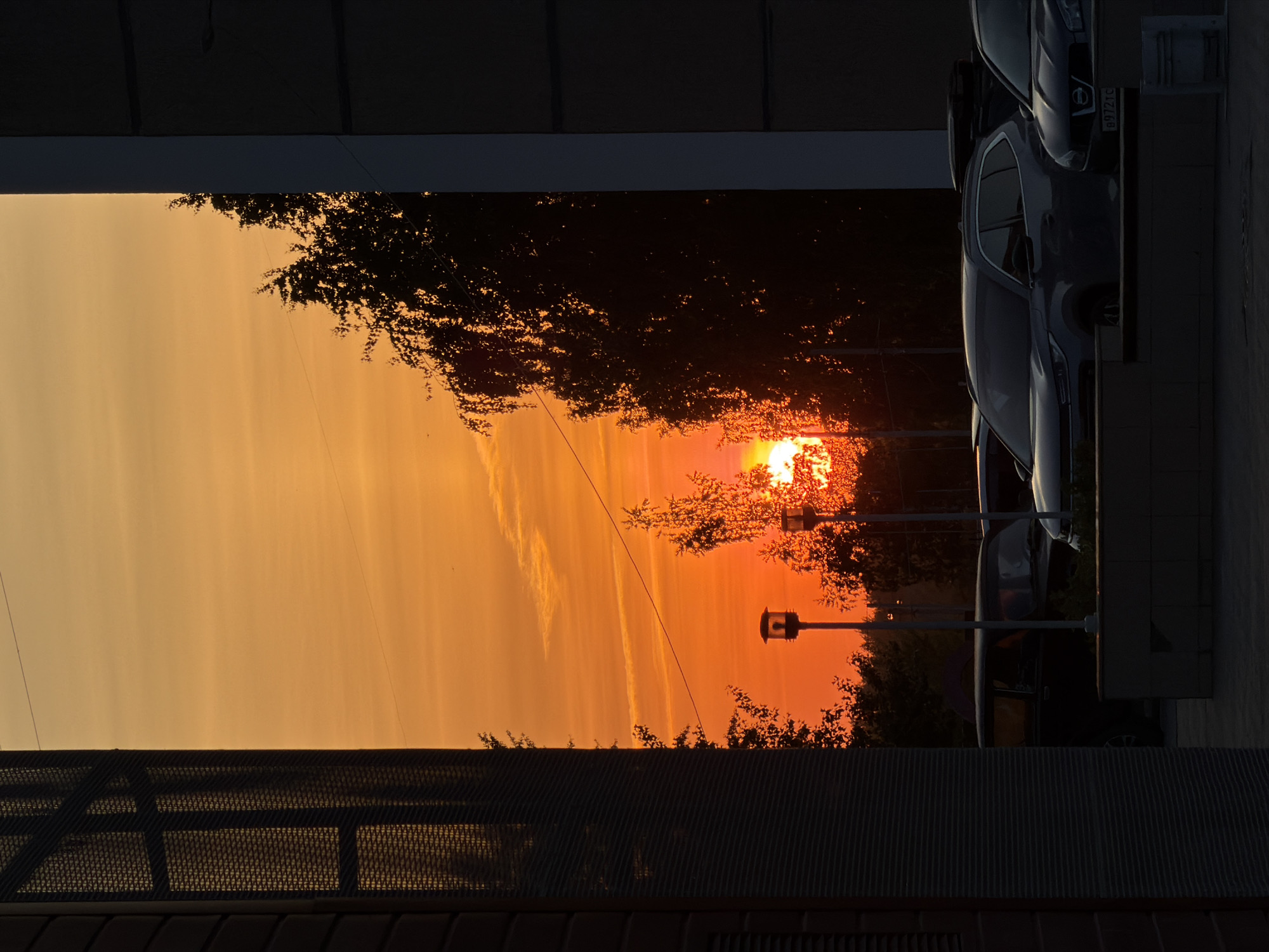 The image depicts a vibrant sunset in Chaikovsky, Russia, viewed between buildings and foliage. The sun is setting behind trees, casting an orange glow across the sky. Several parked cars are visible on the right side of the frame. The foreground features architectural details, including a fence. The scene appears to be taken from an elevated position, likely a window or balcony.