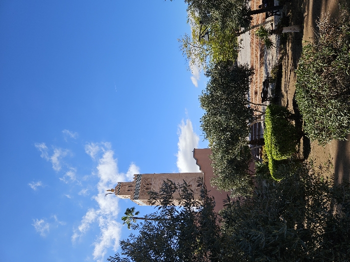 A tall minaret rises against a vibrant blue sky dotted with fluffy white clouds. Lush green trees and bushes surround its base, partially obscuring its lower levels. Sunlight casts shadows across the manicured garden, highlighting a brick walkway and benches. The minaret's intricate design adds a touch of traditional architecture to the scene.