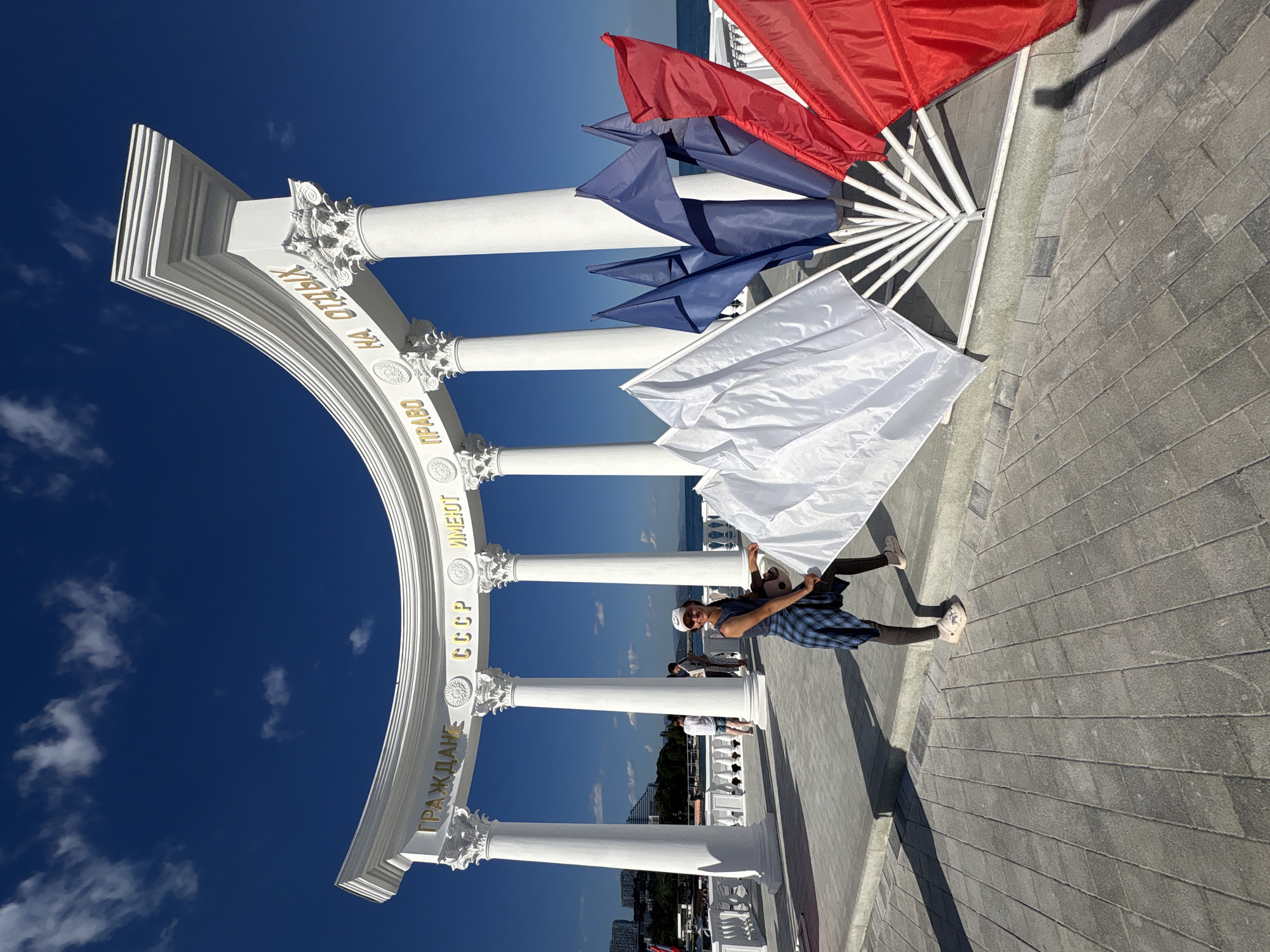 The image shows a woman standing in front of a white, classical-style archway with columns. The archway has text written on it in gold letters. To the right of the woman are several flags, some white, some blue, and some red, arranged in a fan shape. The woman is wearing a blue plaid shirt, dark leggings, and white sandals. She is holding a large white flag. In the background, there is a body of water and buildings. The sky is blue with some white clouds. The text on the archway is in Cyrillic script and reads "ГРАЖДАНЕ СССР ИМЕЮТ ПРАВО НА ОТДЫХ", which translates to "Citizens of the USSR have the right to rest." This monument is located in the city of Alushta, Ukraine.