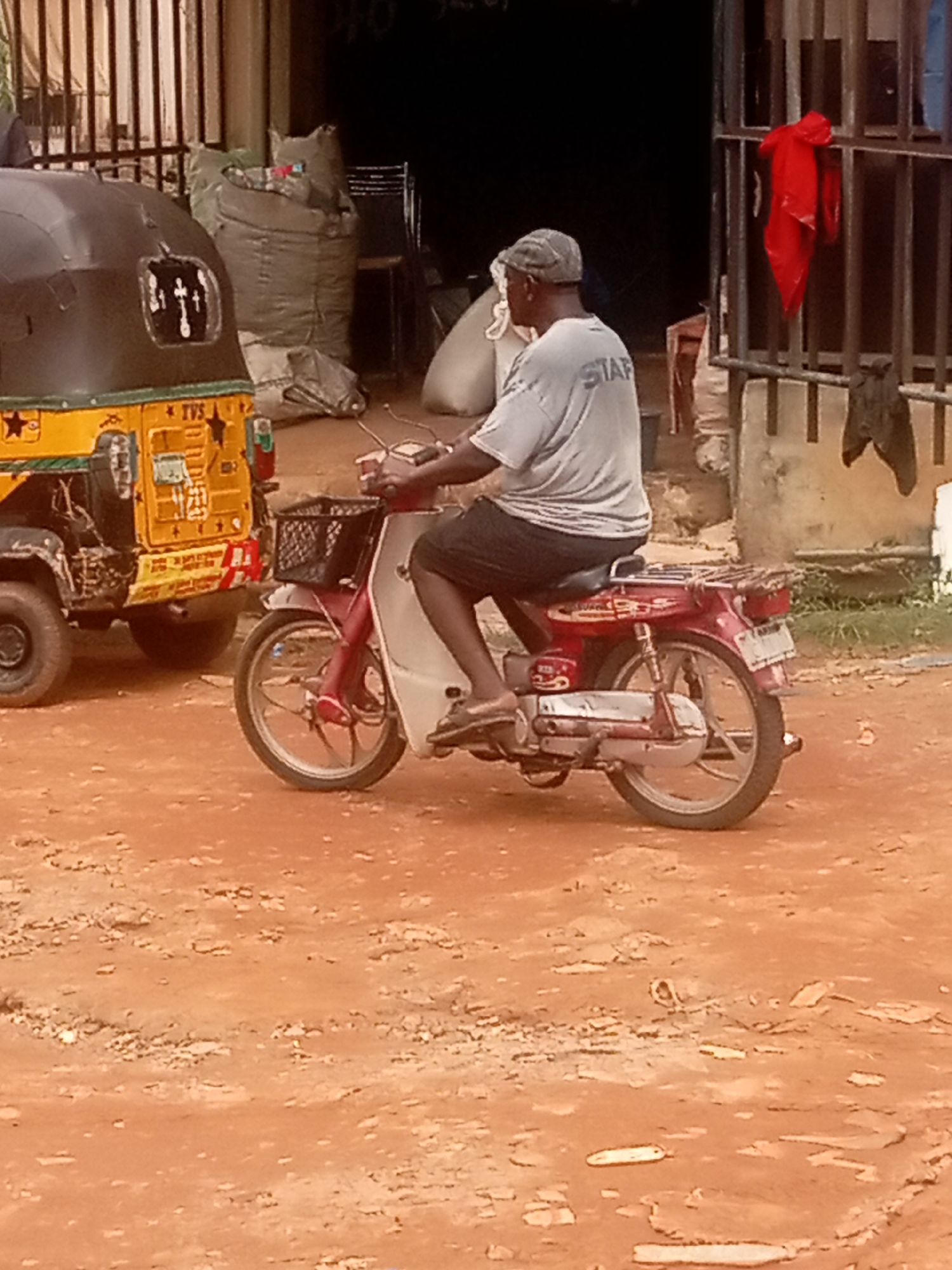 The image shows a man riding a red and white scooter in Umuokpu, Nigeria. He is wearing a light gray shirt with the word "STAFF" printed on the back, black shorts, and a gray hat. He is wearing brown sandals. There is a yellow tuk-tuk parked to the left of the scooter. The ground is red dirt. In the background, there is a building with a metal gate and bags of some sort. A red piece of cloth is draped over the gate.