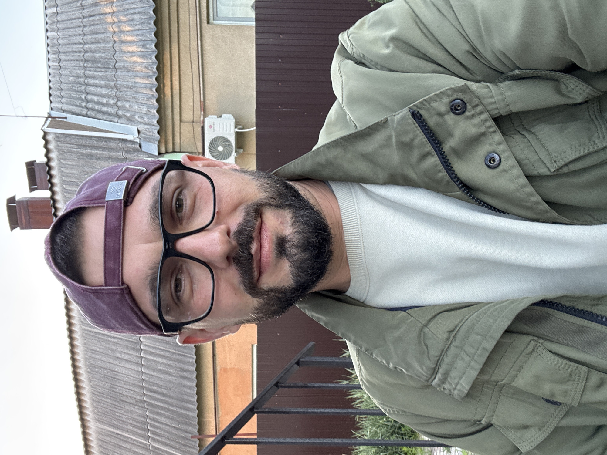 This is a portrait-style selfie of a man in Novaya Usman', Russia. He is wearing a green jacket over a white sweater and a purple baseball cap. He also has black-rimmed glasses and a beard. The background suggests an outdoor setting, possibly a backyard or a street, with a house, a corrugated roof, and a metal fence visible. An air conditioning unit is mounted on the exterior wall of the house. The lighting suggests it is daytime, likely late afternoon or early evening. The man's expression is neutral. The visible text on the hat reads "max&co".