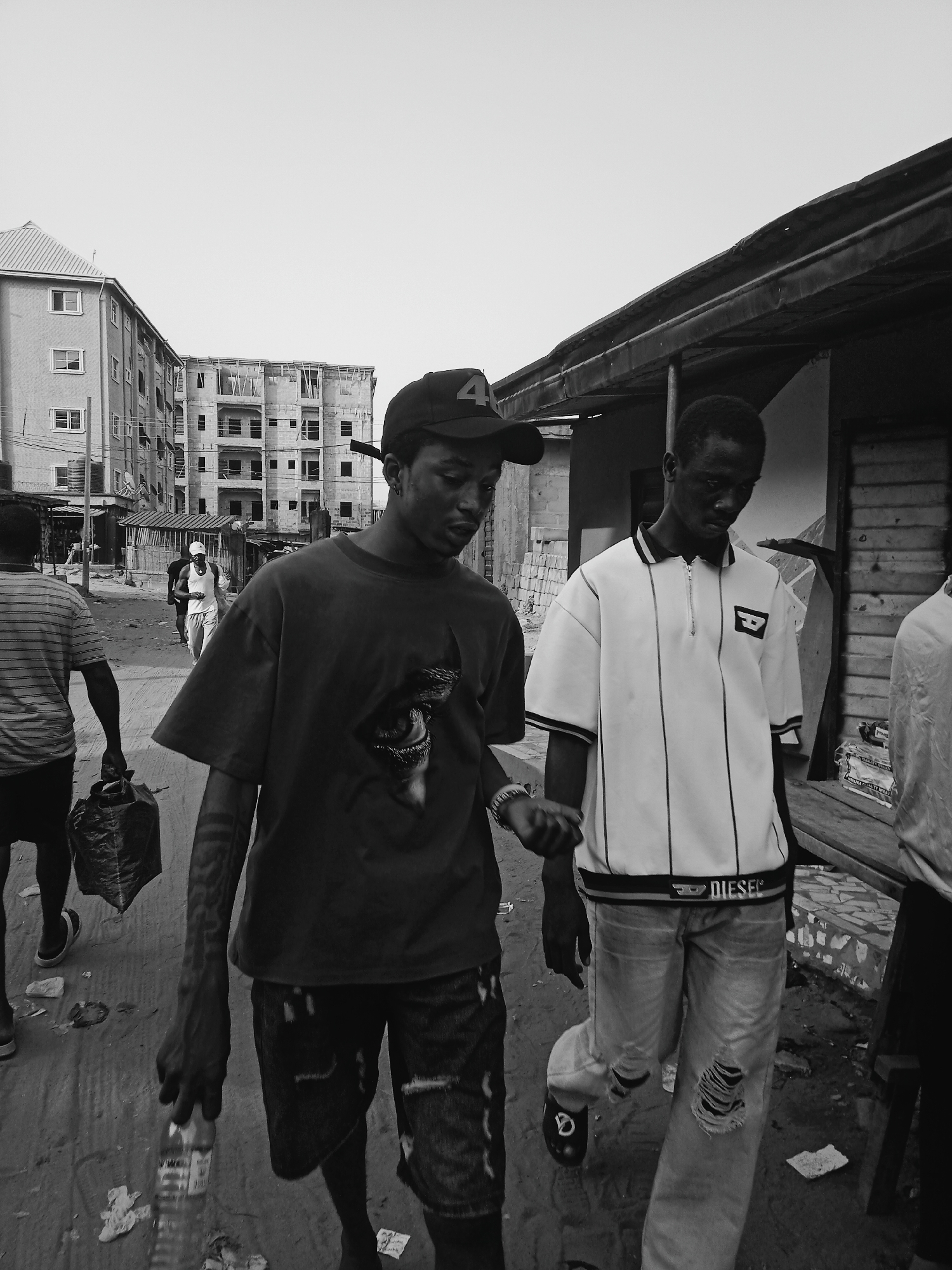 The image, in black and white, shows a street scene in Onitsha, Nigeria. Two young men are walking down a street. The man on the left wears a dark T-shirt, a baseball cap with the number '4' on it, and patterned shorts. He carries a plastic bottle in his left hand. The man on the right wears a white, striped polo shirt, ripped jeans, and slip-on shoes. In the background, there is a man in a striped shirt carrying a black bag. Further back, a woman in white clothing walks down the street. Buildings are visible in the background, including a multi-story building that appears to be under construction. The environment looks urban. The ground is dirt and uneven.