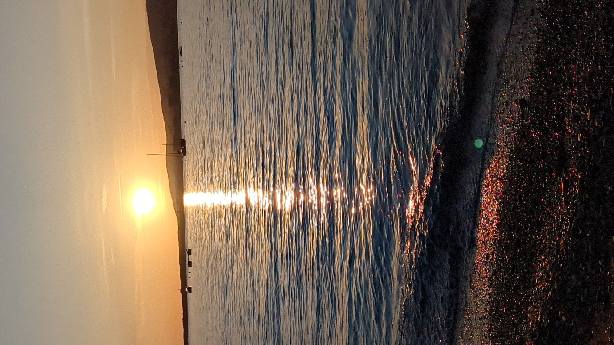 The image depicts a serene coastal landscape during either sunrise or sunset, characterized by a warm, golden light. A bright, glowing sun is positioned low on the horizon, casting a brilliant light that creates a dazzling path of shimmering reflections across the water surface.
In the distance, a dark landmass, likely hills or mountains, is silhouetted against the hazy, luminous sky. Faint, dark shapes, possibly small boats or buoys, are visible on the water near this distant land.
The right side of the frame shows the shoreline where the water meets a dark, pebbly or sandy beach. The wet ground of the beach sparkles with numerous tiny, colorful reflections of the sun's light, creating a glittery effect. A notable detail is a distinct green circular lens flare visible in the water near the shoreline. The water surface itself is textured with subtle ripples, further enhancing the play of light.
The scene conveys a peaceful and picturesque mood, showcasing the natural beauty of the sun interacting with the sea and shore. The location context indicates this tranquil scene is in Gelendzhik, Russia. There is no visible text in the image.