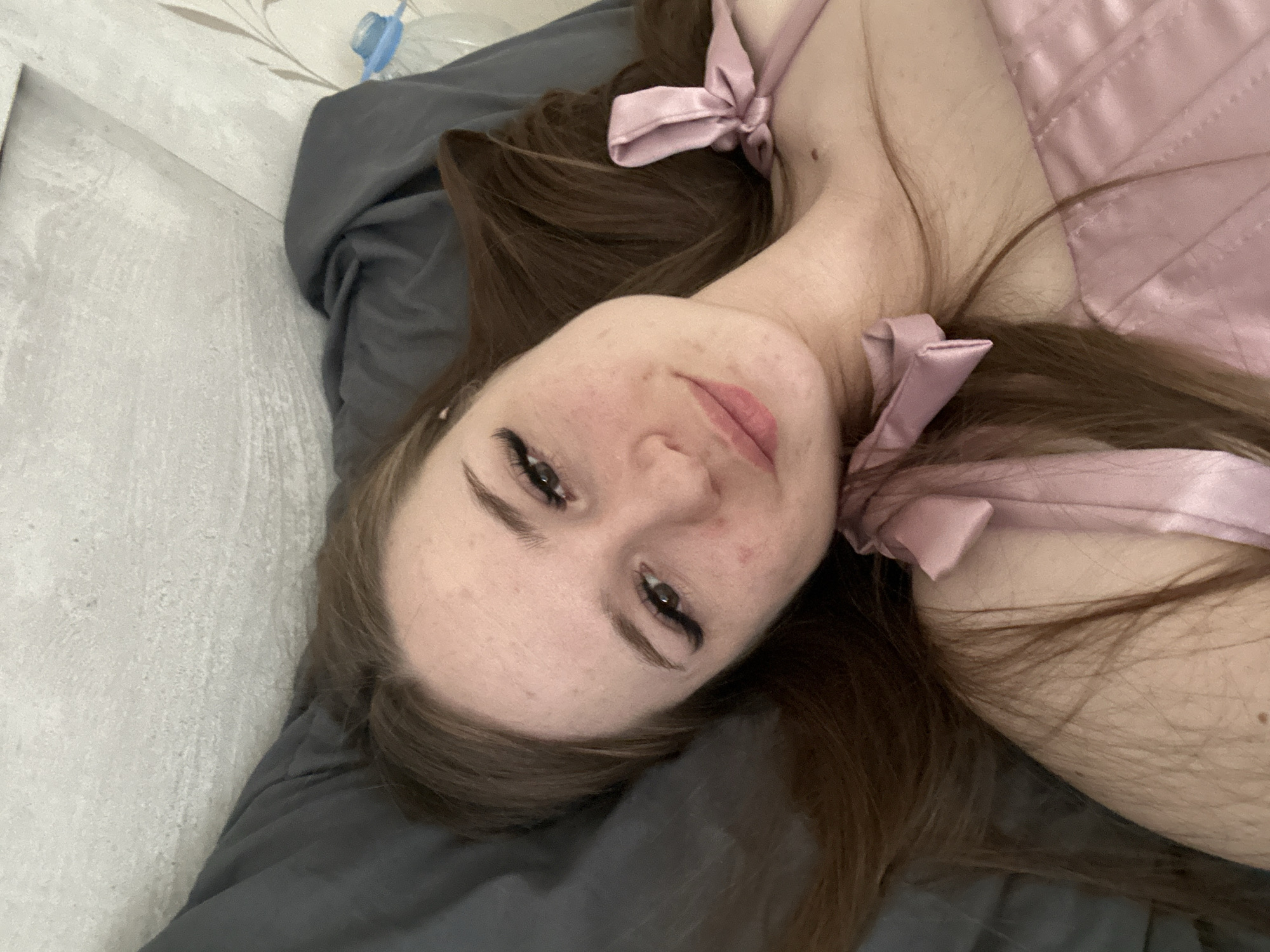 A person lies on a gray pillow wearing a pink, satin top with bows. The person has long, brown hair and is looking at the camera. The background includes a wall with a floral design. A water bottle is partially visible. The location is unknown.