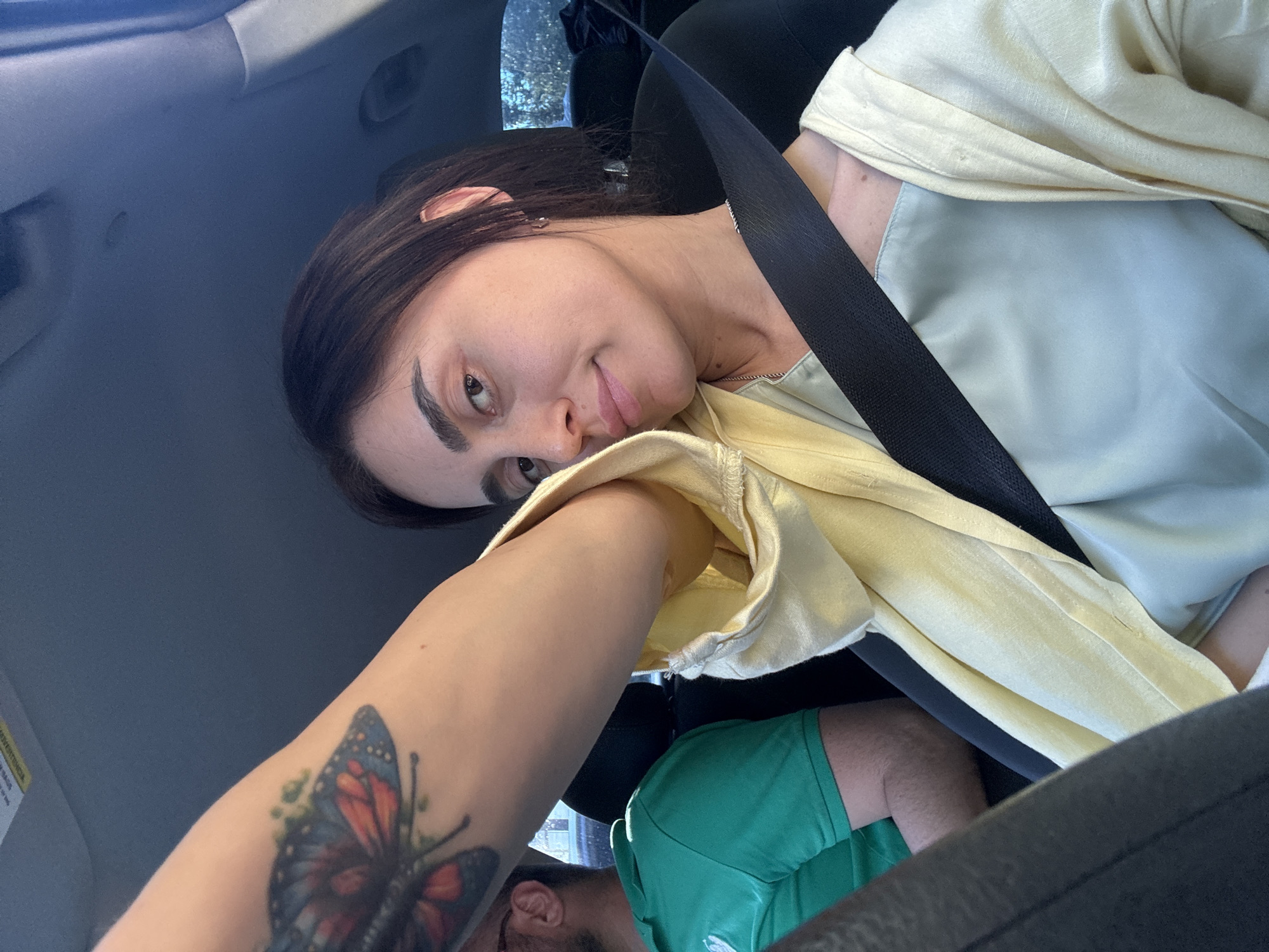 This media file depicts two people inside a vehicle, likely a car, based on the visible seatbelt and interior elements. The primary subject, positioned in the foreground and slightly out of focus, is a person with dark hair, wearing a light yellow or cream-colored garment. Their face is turned towards the camera, with a subtle, perhaps amused or thoughtful, expression. An arm with a butterfly tattoo is extended, partially obscuring the view of the person's torso and the car's interior.
Behind and to the side of the main subject, another person is visible. They are wearing a bright green t-shirt and their arm is resting on their leg. The setting appears to be daytime, with natural light filtering into the vehicle. The presence of trees visible through the windows suggests an outdoor location. The people are seated, and the angle of the shot implies it might be a selfie taken by the person in the foreground, or by someone next to them.
There is a black seatbelt clearly visible, diagonally crossing the frame and securing the primary subject. A yellow tag with some text is visible on the left side of the image, but the text is too blurry to decipher. The overall mood seems casual and relaxed. Given the provided location context of Tbilisi, Georgia, the scene is situated within that city.