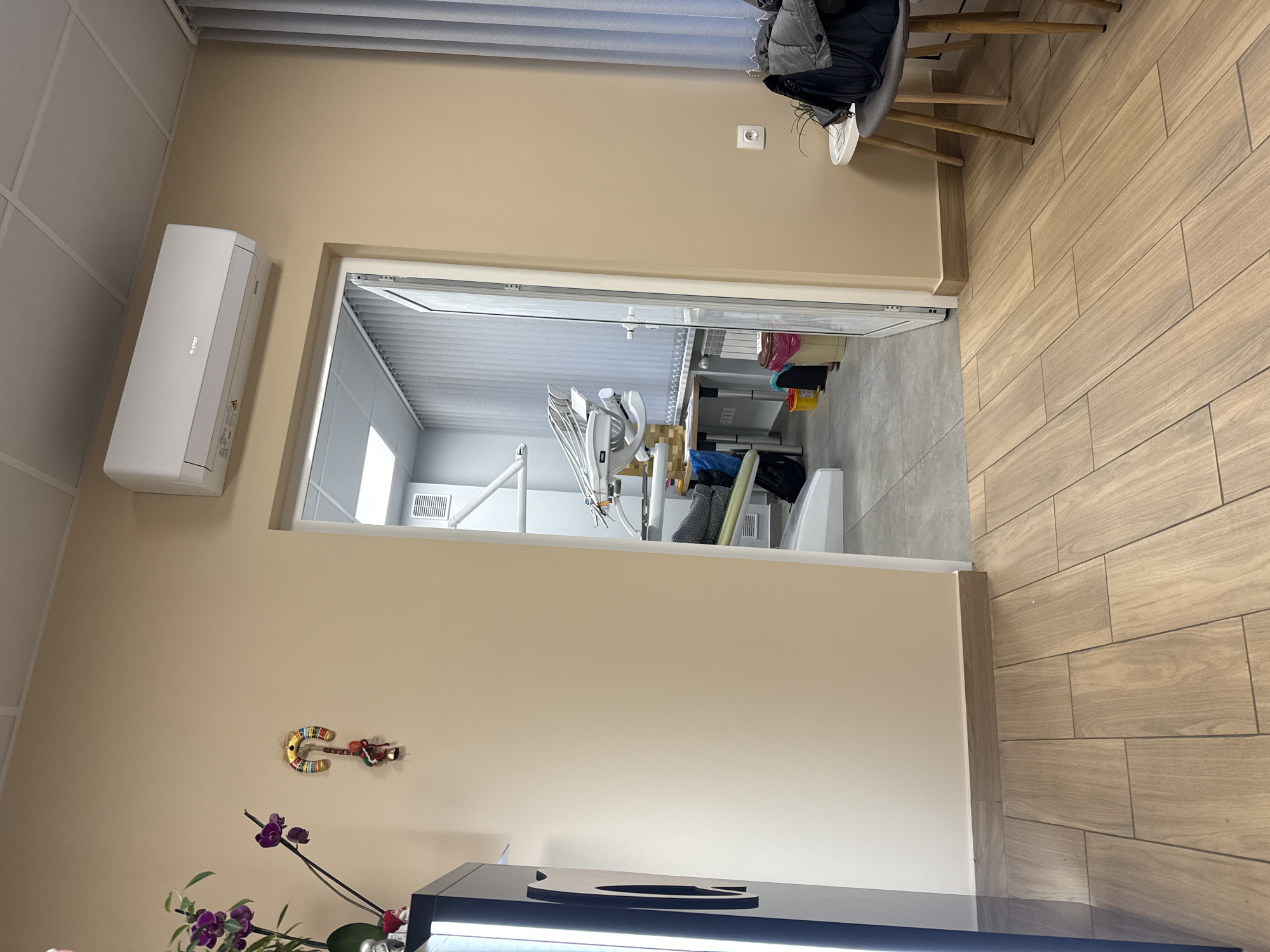 The interior of what appears to be a dental office or clinic is captured in this shot. The walls are painted a light beige color. Visible near the top of the frame, there's an air conditioning unit mounted on the wall. Below it is an open doorway leading into a brighter, sterile-looking room containing dental equipment, including a dental chair and various apparatus. On the wall to the left is an ornamental art piece and below it a small plant. The flooring on the right is wood-grain tiling. A chair with clothing draped over it is partially visible in the upper right corner of the frame. The location appears to be in Sofia, Bulgaria.