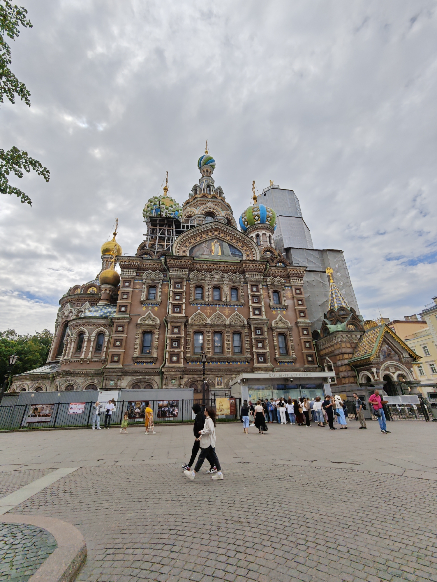 The image captures a low-angle, wide-angle view of the Church of the Savior on Spilled Blood in Saint Petersburg, Russia, under a heavily overcast sky. The distinctive onion domes of the church, some adorned with green, blue, and gold mosaics, are partially obscured by scaffolding and a large grey tarp covering a section of the building, indicating ongoing construction or renovation. The intricate facade of the church, featuring colorful ceramic tiles and ornate architectural details, is visible.
In the foreground, a paved cobblestone plaza is occupied by a diverse group of people. Several individuals are walking across the plaza, including a pair in the immediate foreground dressed in dark and light casual clothing, with one person in black and white athletic wear and the other in a white top and dark pants. Further back, a line of tourists waits outside a building labeled "КАССЫ TICKET OFFICE." Other individuals are seen standing and conversing in small groups. Fencing with informational posters is set up in front of the church. Trees and parts of adjacent buildings can be seen to the left and right of the church, respectively, suggesting an urban setting. The overall atmosphere is one of a popular tourist destination experiencing overcast weather, with ongoing restoration work on a prominent landmark.