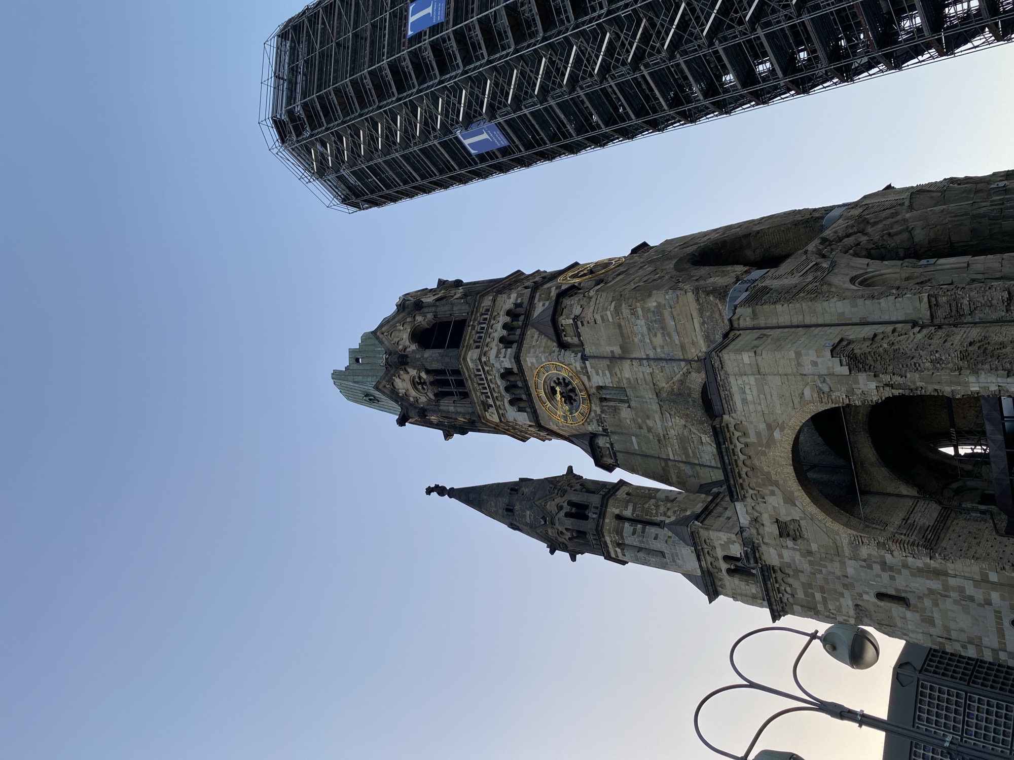 The image shows a view of the Kaiser Wilhelm Memorial Church in Berlin, Germany. The church's damaged spire and remaining structure are visible. The sky is clear and light blue. A large metal scaffolding structure is situated above the church. A street lamp and possibly some panels are in the foreground.