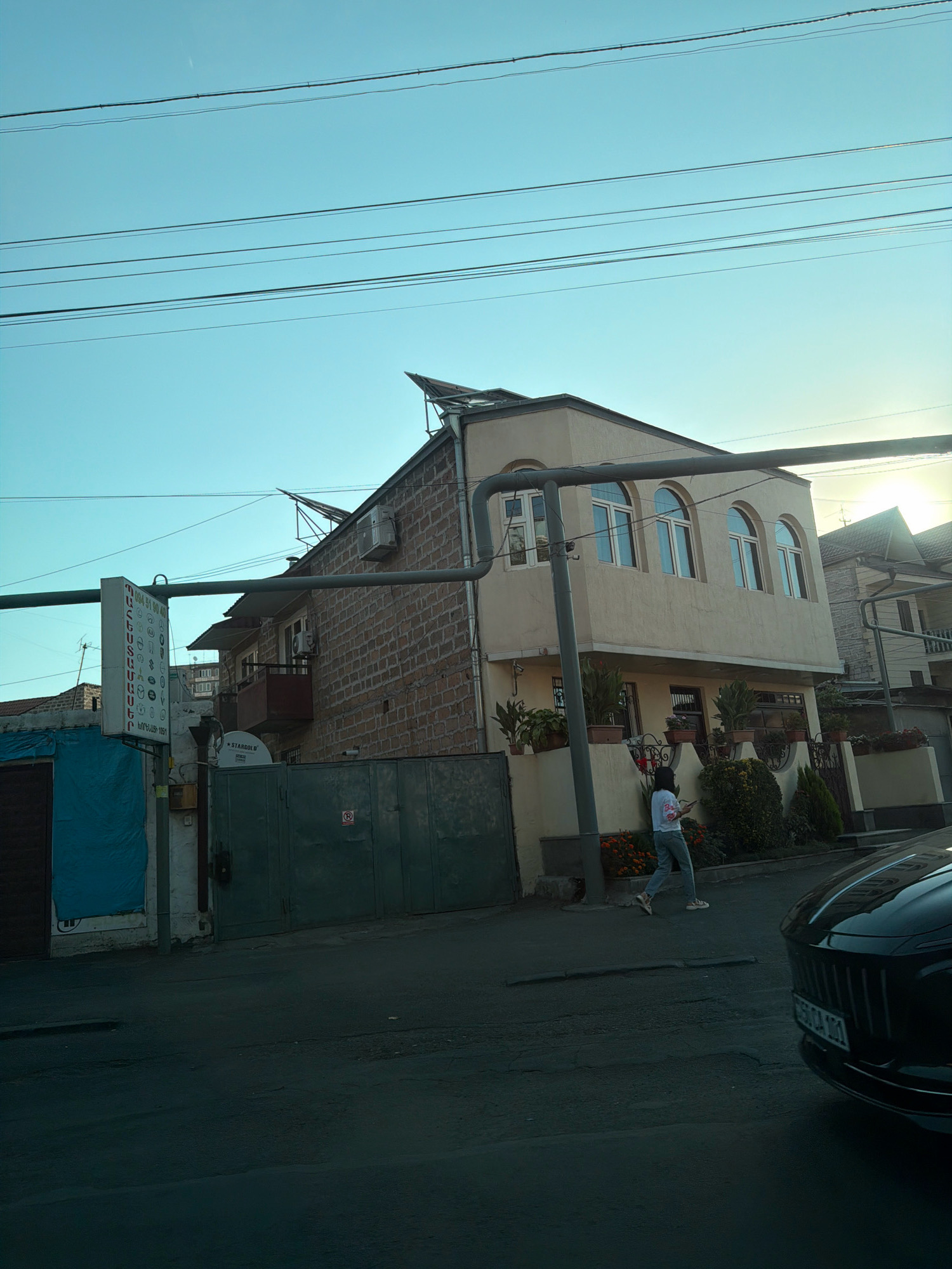 In Yerevan, Armenia, a scene unfolds with a two-story building featuring arched windows and a balcony. A person in white shirt and grey pants walks on the sidewalk. Adjacent to the building, a metal gate and a blue tarp-covered structure are visible. Overhead, power lines crisscross the clear blue sky. A black car is parked in the foreground.
