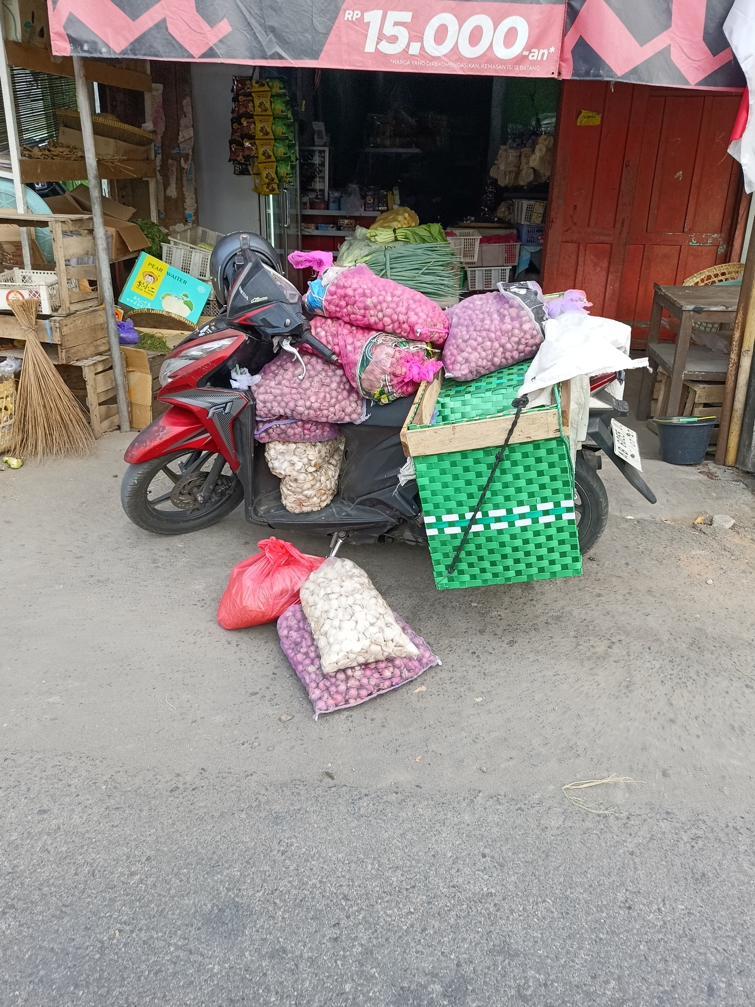 A red Honda scooter is parked on a paved surface, presumably a street or market area, outside a small shop. The scooter is loaded with several large sacks of onions and garlic. Two of the sacks are red and purple net bags filled with onions and are placed on the seat. A green plastic woven basket is attached to the rear of the scooter, also holding sacks of onions and a white plastic bag. More sacks of produce, including what appears to be garlic and more onions, are piled on the ground next to the scooter, with a red plastic bag also on the ground. In the background, the shop has a red wooden door and shelves displaying various goods, including snacks and possibly other produce. A sign above the shop entrance displays the text "RP 15.000". A broom and some wooden crates are visible to the left of the scooter. The scene suggests a vendor preparing to transport or sell produce in Sukorini, Indonesia.