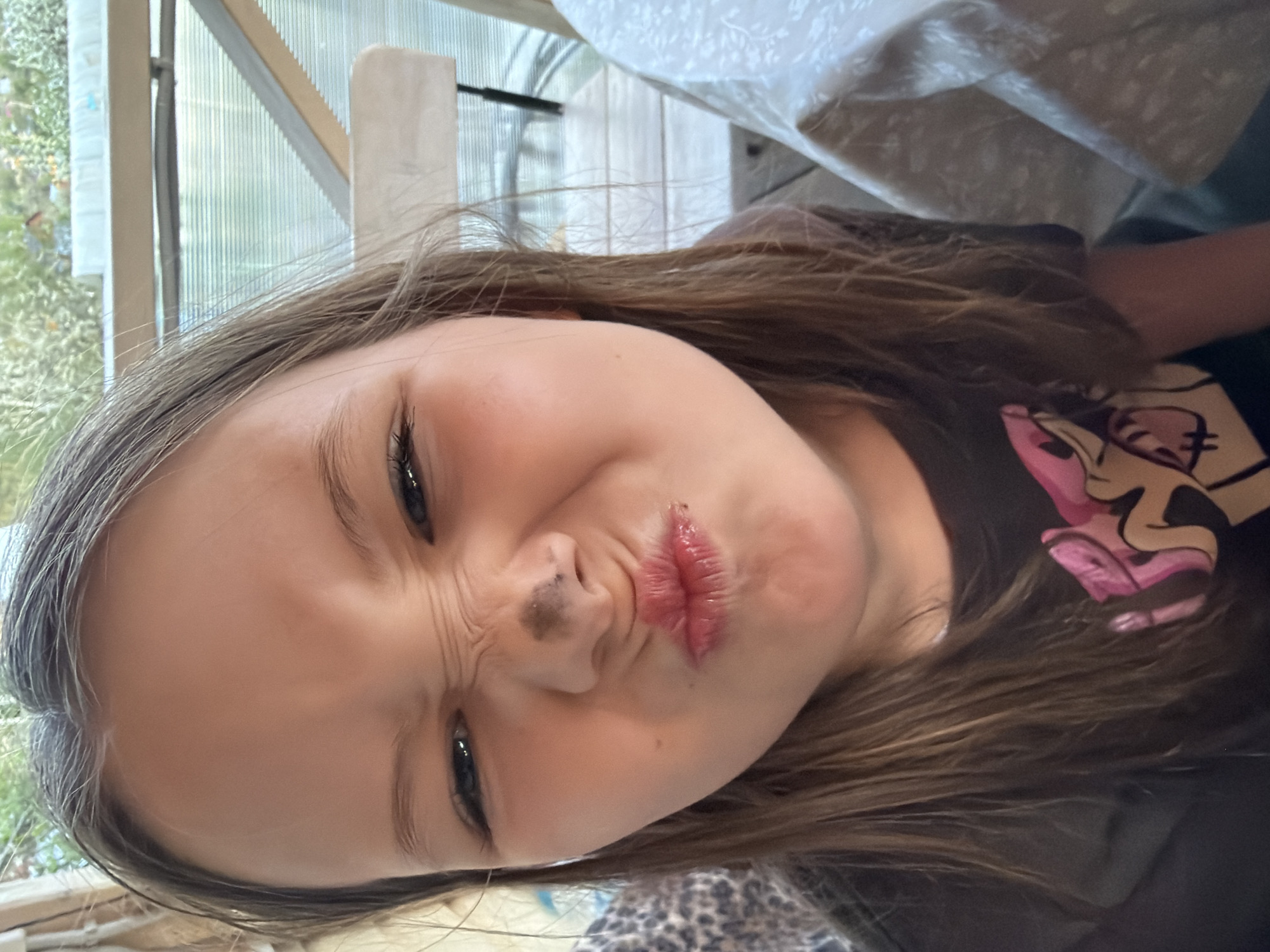 A close-up, rotated image shows a young girl with shoulder-length brown hair. She is making a pouting face with pursed lips and wrinkled nose. There is a dark smudge on her nose. She is wearing a dark t-shirt with a cartoon graphic featuring pink. The background appears to be a room with a window or screened-in area to the left, possibly a greenhouse. Part of a white tablecloth is visible above her head. No definitive evidence to verify the location as Chaikovsky, Russia, is discernible in the provided image.