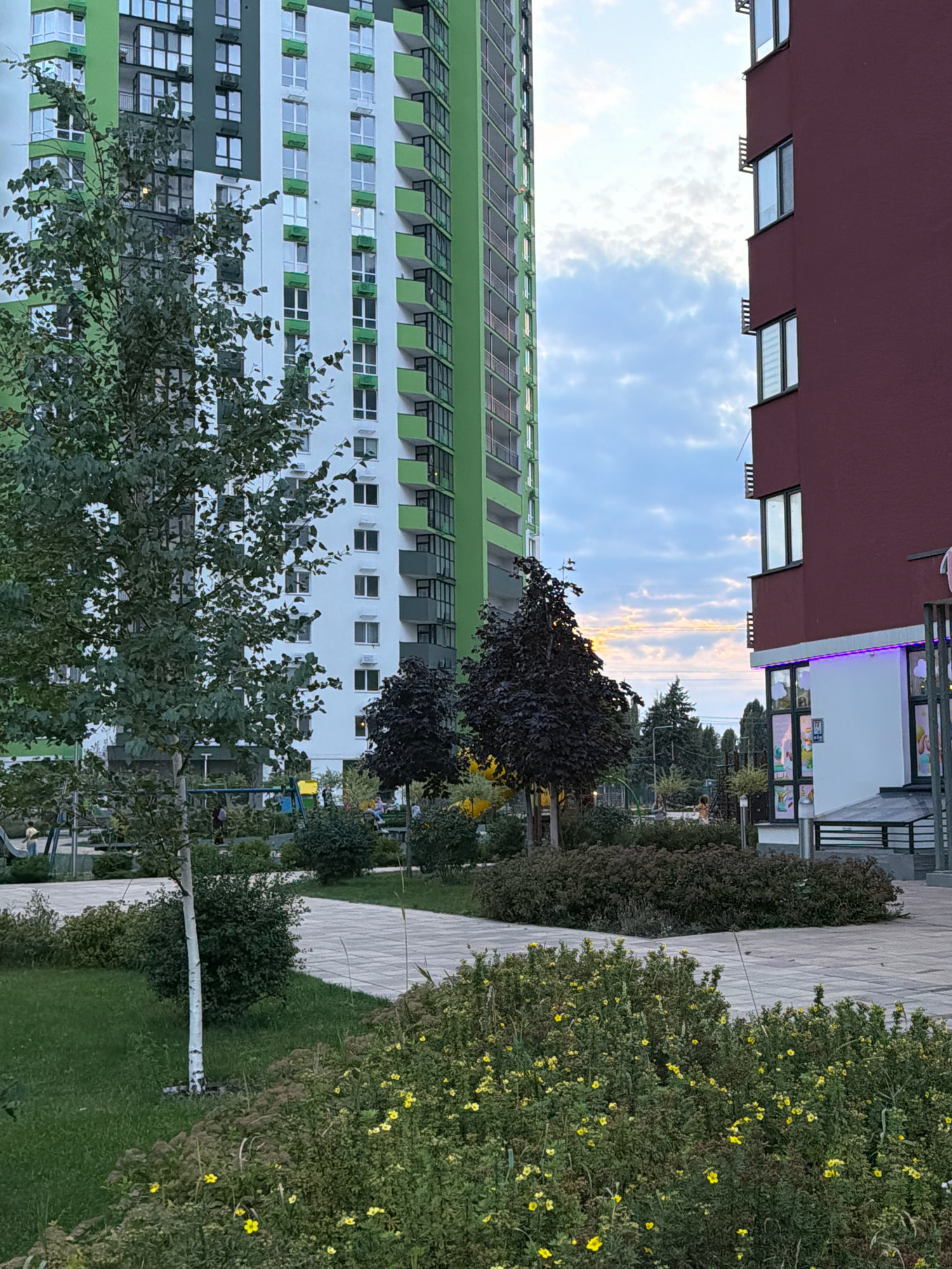 The image depicts a residential complex in Kyiv, Ukraine, characterized by two high-rise buildings. The building on the left is predominantly white with green accents, featuring numerous windows and balconies. To its right, a smaller building with a maroon facade is visible, also with multiple windows. The foreground shows a well-maintained outdoor area with paved walkways, lush green grass, and abundant vegetation, including bushes with small yellow flowers and trees with dark foliage. In the background, a playground with colorful equipment can be partially seen. Several individuals are present in the scene, including some sitting on benches and others near the playground. The sky above is partly cloudy with hints of sunset colors. The overall impression is that of a modern urban residential environment designed with green spaces and recreational facilities.