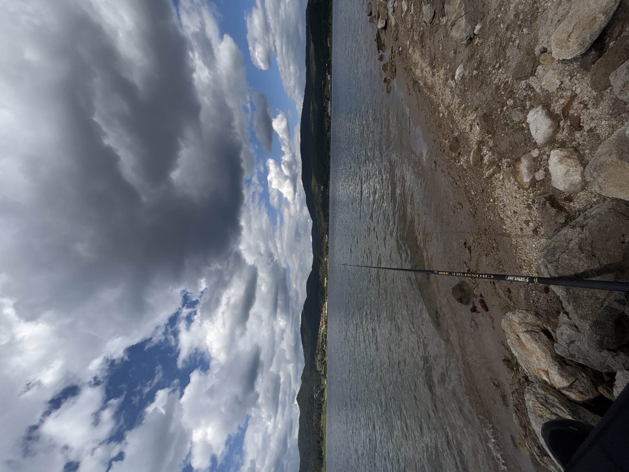 A fishing rod stands upright on a rocky shore, its tip angled towards a large body of water under a dramatic sky filled with dark, heavy clouds interspersed with patches of bright blue. The water is rippled, and a distant shoreline is visible, featuring a dense, dark green forest that climbs the rolling hills. A small cluster of buildings, appearing red-roofed, is nestled among the trees on the hillside. The foreground is dominated by various-sized rocks and a sandy, pebbly beach. The image is shot from a low angle, focusing on the fishing rod and the immediate shoreline.