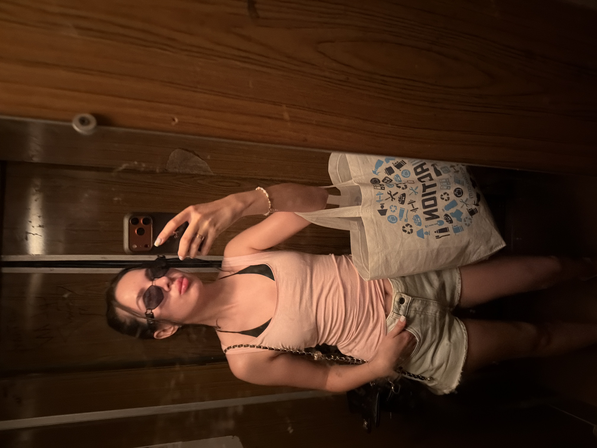 A young woman is taking a mirror selfie in what appears to be an elevator. She is wearing a light pink tank top, a black bikini top underneath, and light-colored denim shorts. She has dark hair pulled back, dark sunglasses, and is holding her phone in her right hand, with her left hand in her pocket. She is also carrying a white canvas tote bag with various icons printed on it. The elevator walls are wood-paneled and show some signs of wear.