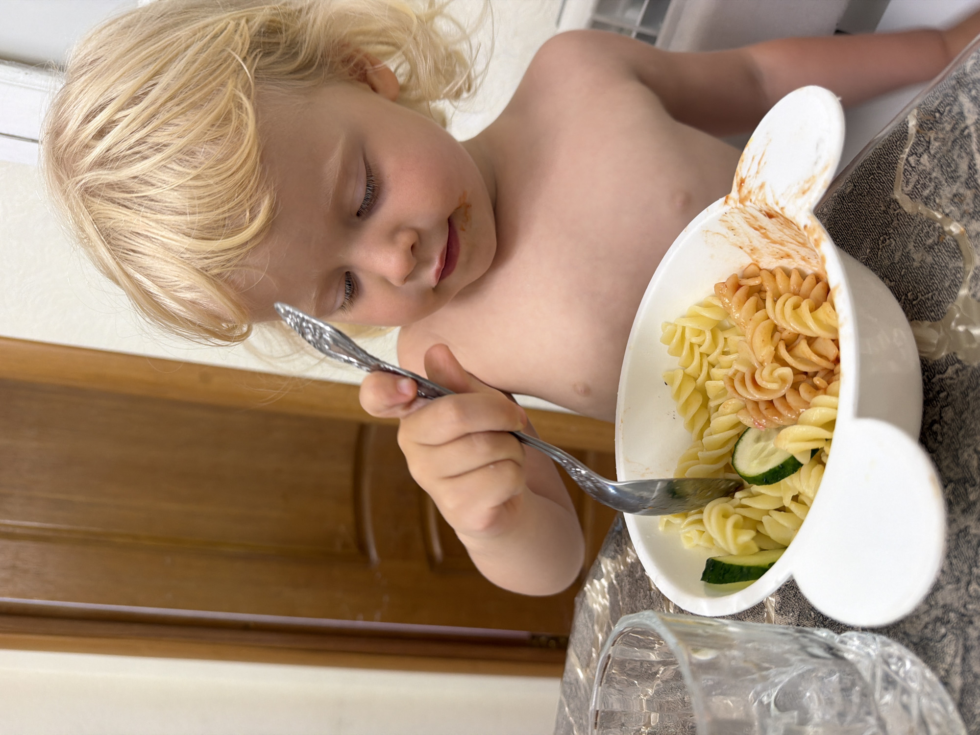 This is a close-up, eye-level shot of a young child with blonde hair eating pasta. The child, who appears to be a toddler, is shirtless and leaning towards a white bowl filled with fusilli pasta and slices of zucchini. A metal fork, held by the child's left hand, is partially submerged in the pasta. The child's face is turned downwards, and their eyes are closed, suggesting a moment of focus or contentment while eating. A small smudge of sauce is visible on the child's cheek.
The setting appears to be indoors, possibly a dining area. Behind the child, a wooden surface, possibly a door or furniture, is visible. The table surface in the foreground is patterned and appears to be a dark marble or granite. A clear glass object, possibly a cup or pitcher, is visible in the lower right corner of the frame, reflecting light. The lighting suggests it could be daytime, though no strong indications of time of day or weather are present.
The overall mood of the image is intimate and candid, capturing a simple, everyday moment of a child enjoying a meal. There is no visible text in the image. The location context of Sochi, Russia, does not provide any specific visual cues within this frame.