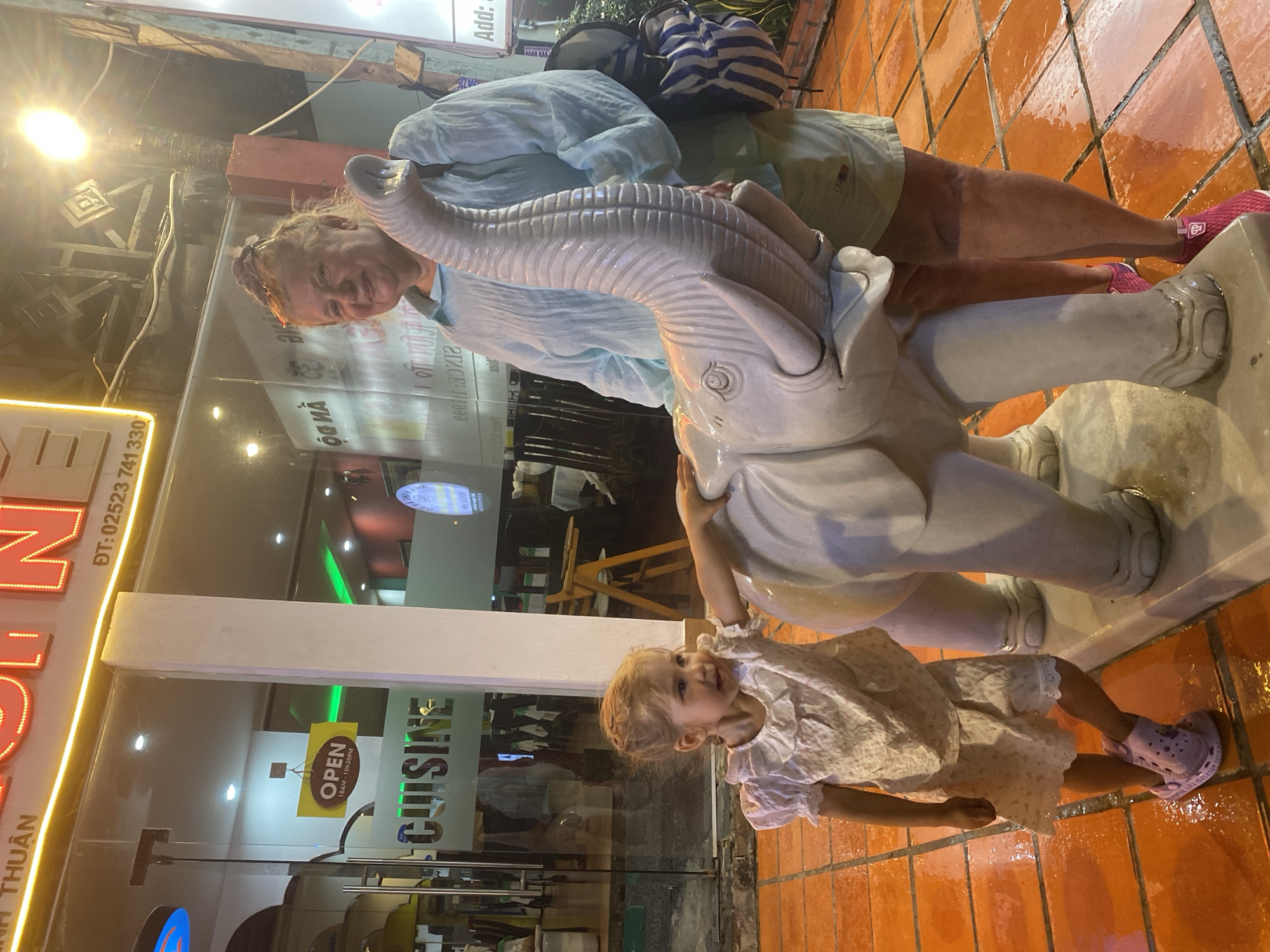 In Mũi Né, Vietnam, a young Caucasian girl in a white floral dress and pink clogs poses with her hand on a white elephant statue. To her right, an adult Caucasian woman in a light blue blouse and khaki shorts smiles, also interacting with the elephant statue. The scene is set in front of a building with a glass facade displaying "CUISINE" and an "OPEN" sign. The ground is paved with orange tiles, appearing wet. Neon signs and streetlights illuminate the outdoor area.