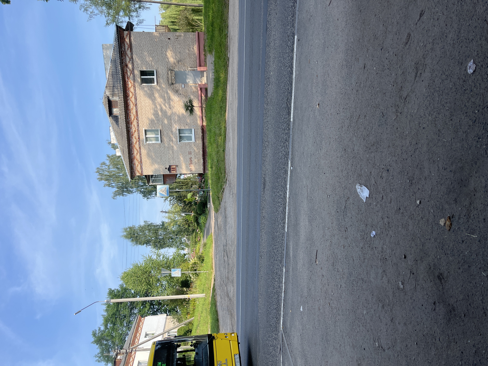 A yellow bus is parked on the side of a road in a suburban or rural area with a two-story brick building in the background. The building has a grey tiled roof and several windows. There are trees and green grass around the building. The road appears to be paved, and there are some white markings on it. The sky is blue with some clouds. The image is taken from a low angle, looking slightly upwards. The license plate of the bus is partially visible. There are two pedestrian crossing signs visible on the side of the road. The overall scene depicts a typical day in a residential area.